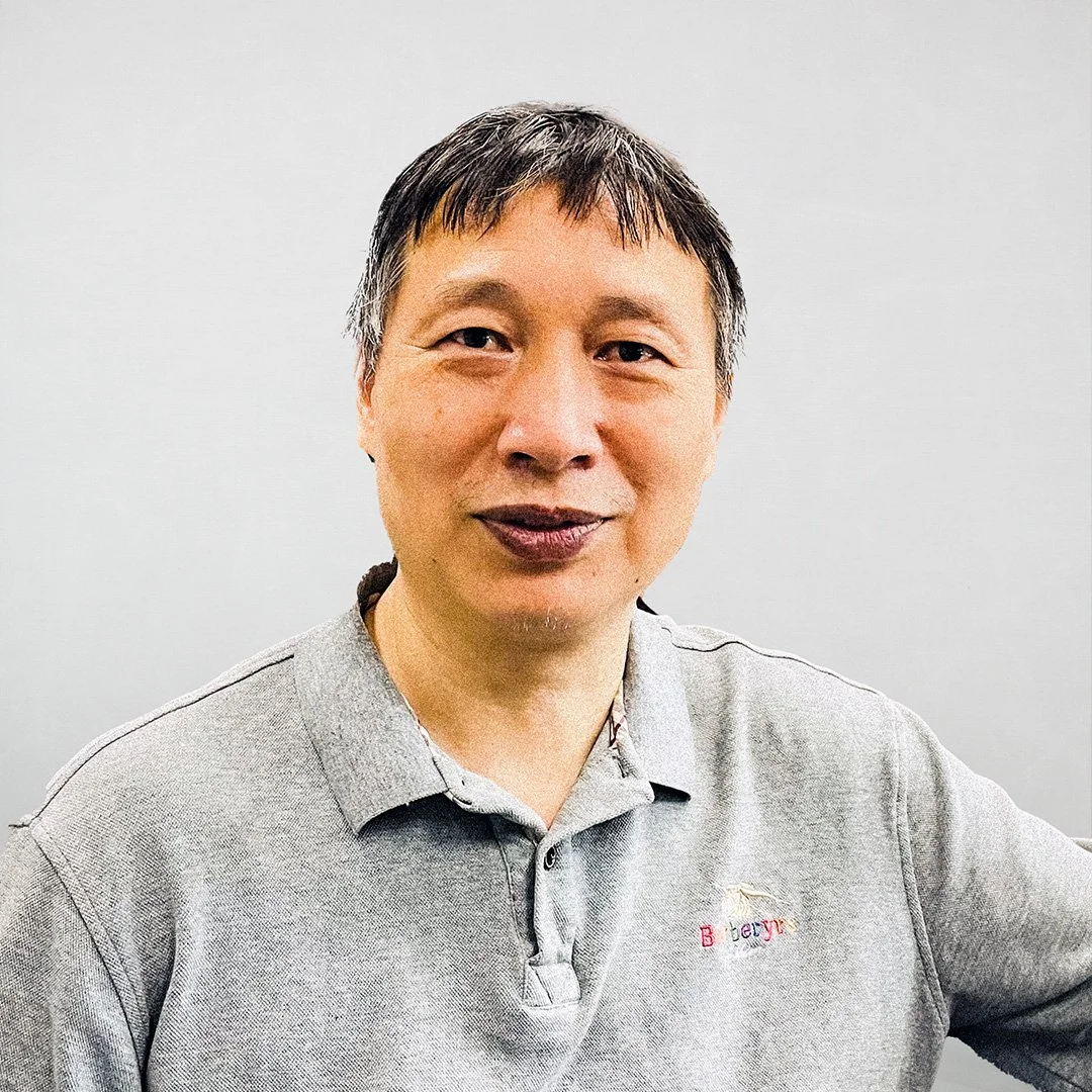 A man with short black and gray hair wearing a gray collared shirt with a small embroidered logo on the chest, posing against a plain light-colored background.