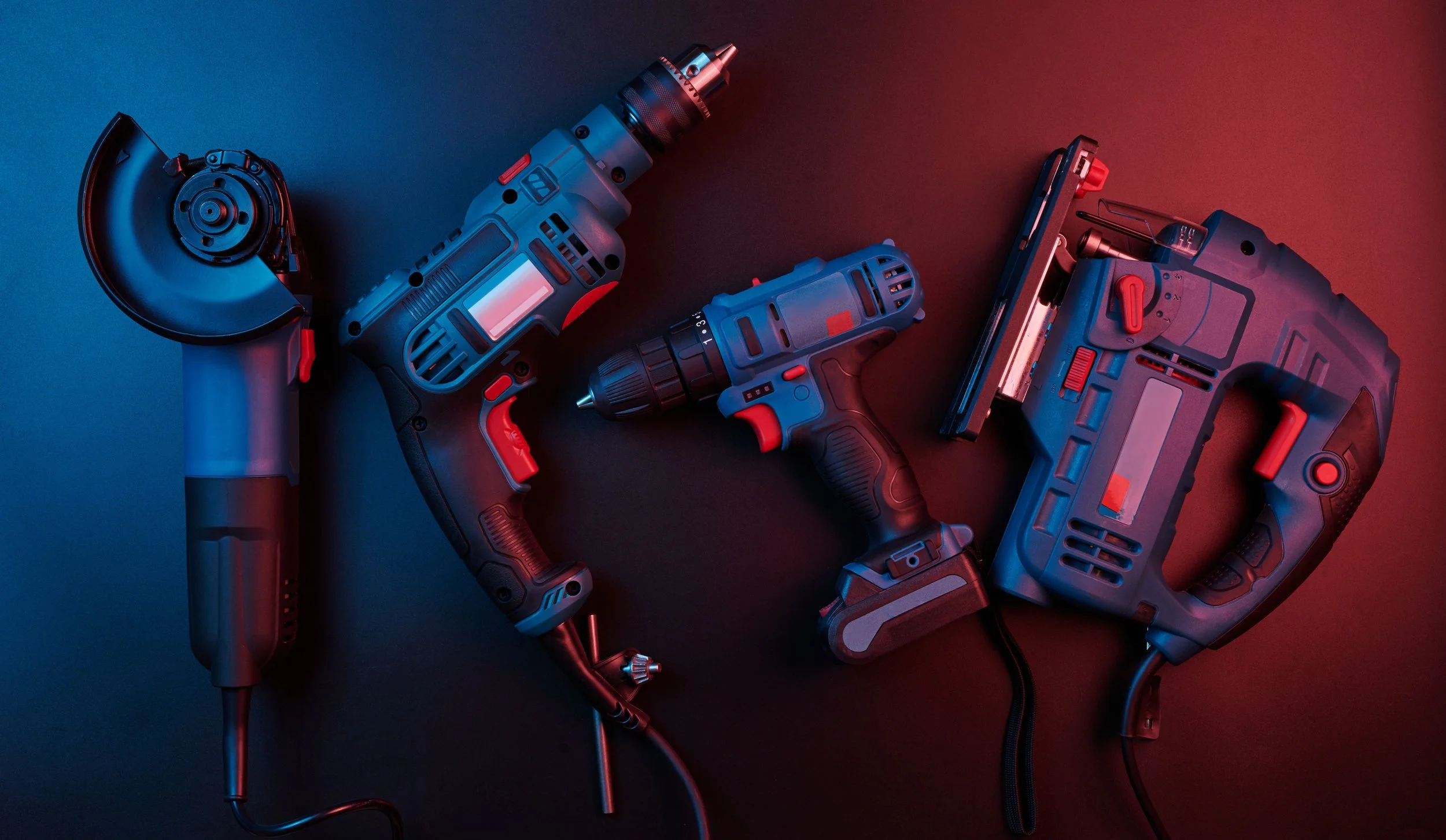 Collection of power tools including a circular saw, cordless drill, impact driver, belt sander, and jigsaw on a surface with blue and red lighting.