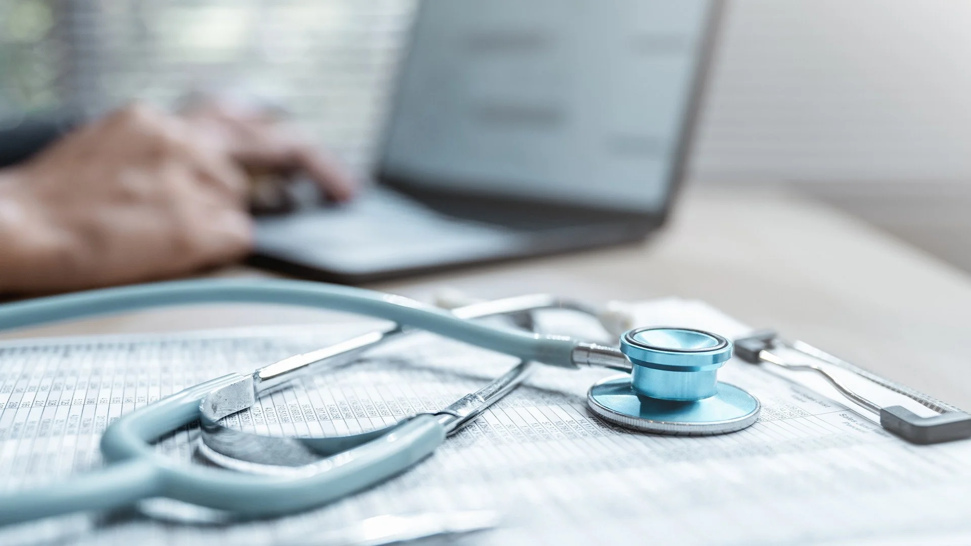 Medical stethoscope placed on a medical chart with a person's hand typing on a laptop in the background.