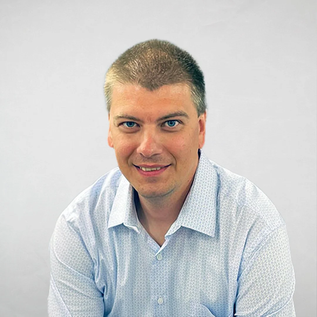 A man with short hair and blue eyes wearing a white collared shirt, smiling at the camera against a plain background.