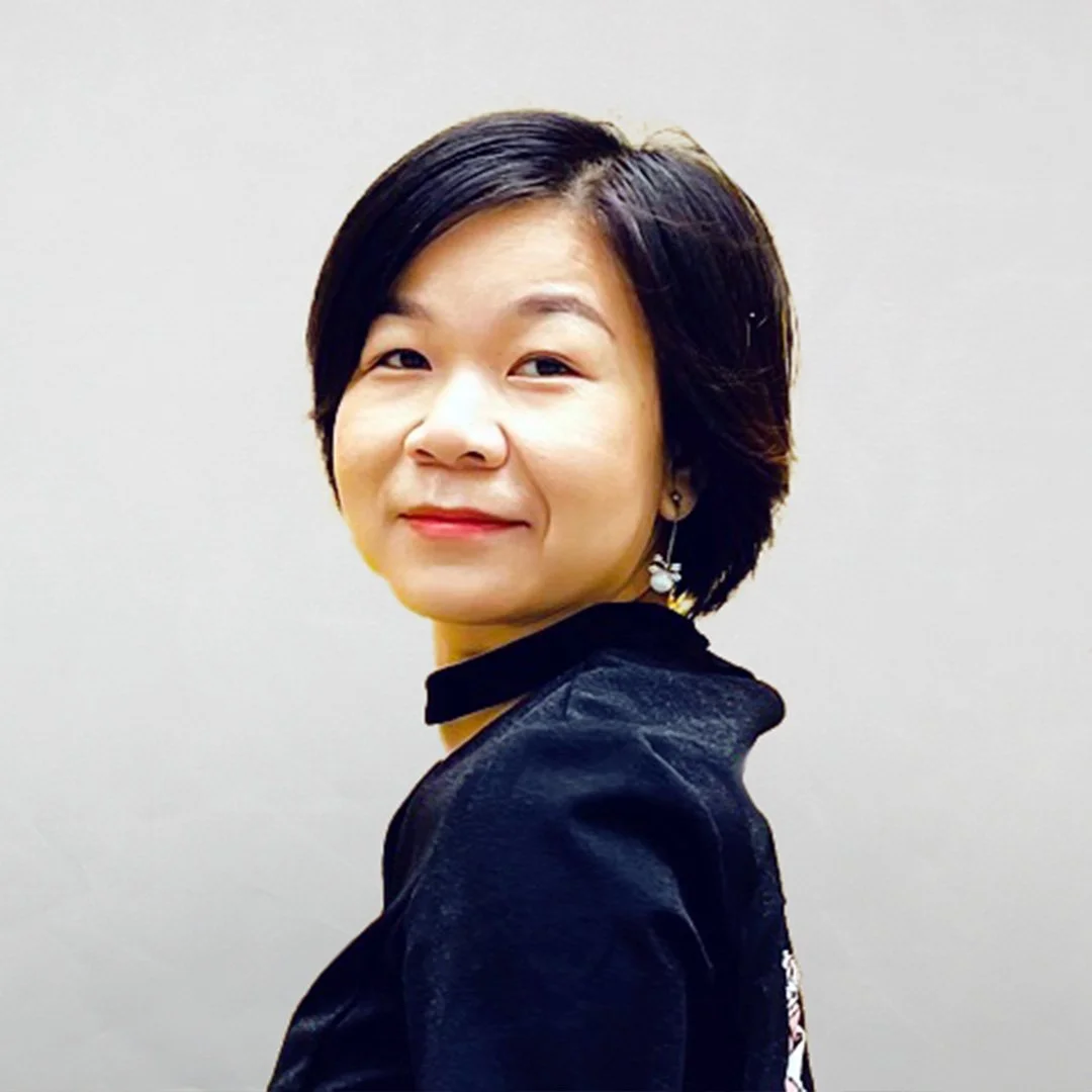 A woman with short dark hair, wearing a black jacket, and earrings, looking at the camera with a slight smile against a plain white wall.