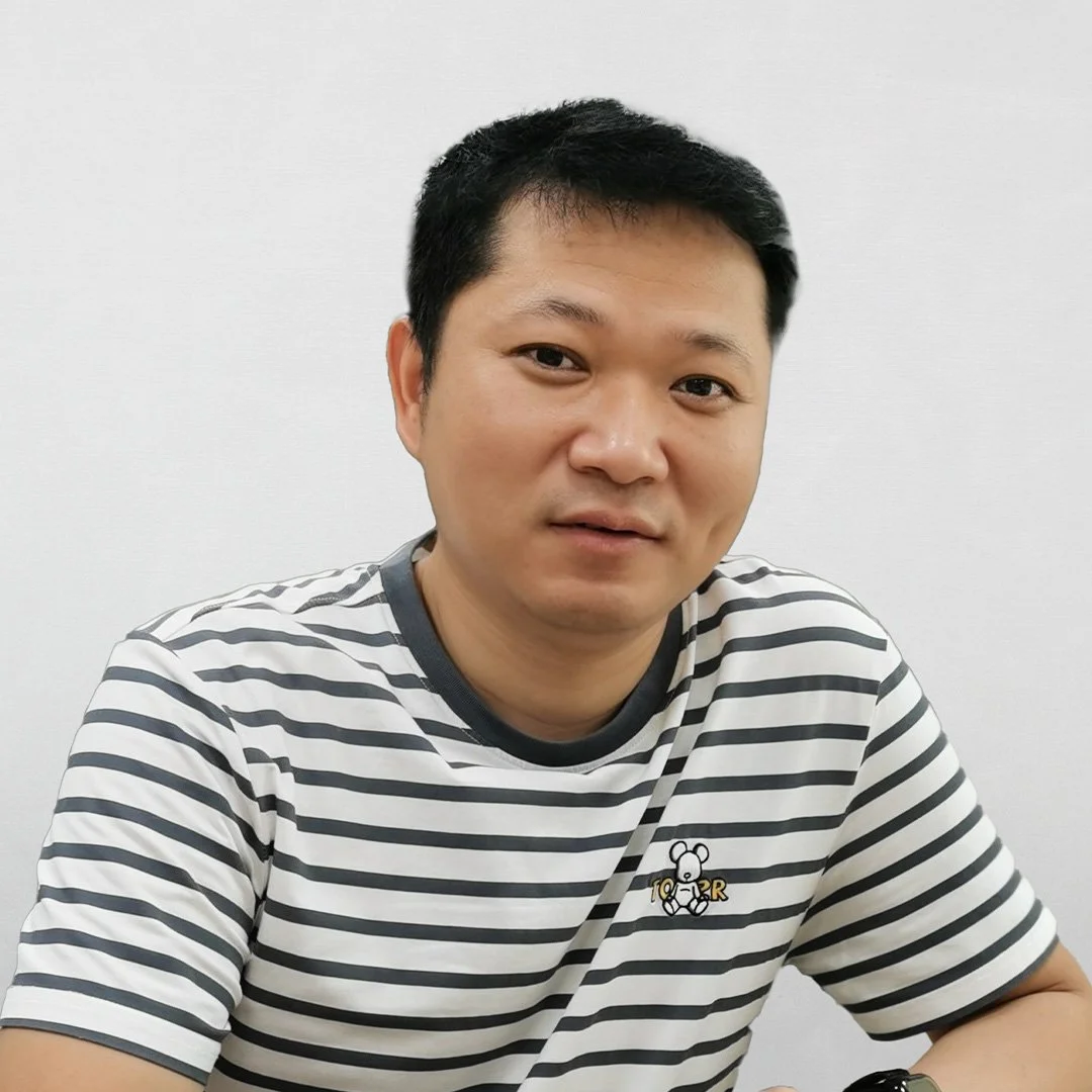 A man with short black hair and a light skin tone, wearing a black and white striped T-shirt with a small bear logo, sitting against a plain white wall.