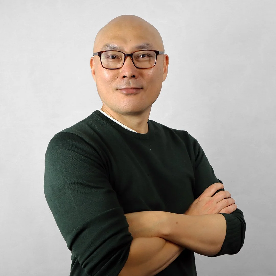 Portrait of a man with glasses, wearing a black shirt, crossing arms, standing against a plain light gray background.