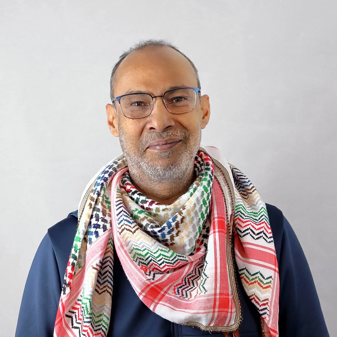 A middle-aged man with glasses, gray beard, and mustache wearing a dark jacket and a colorful patterned scarf against a plain light background.