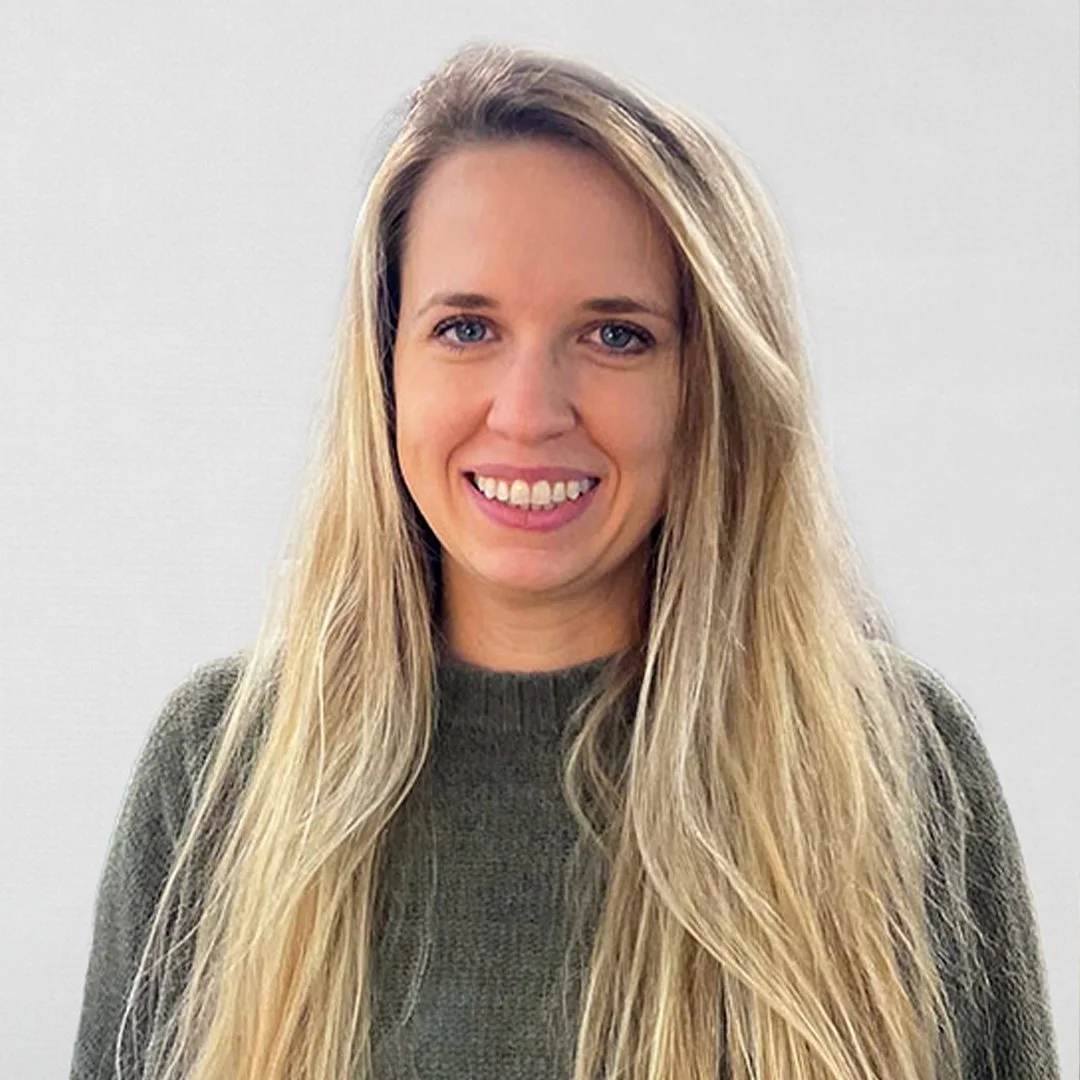 Close-up of a woman with long blonde hair, smiling, wearing a dark green sweater, against a plain light gray background.