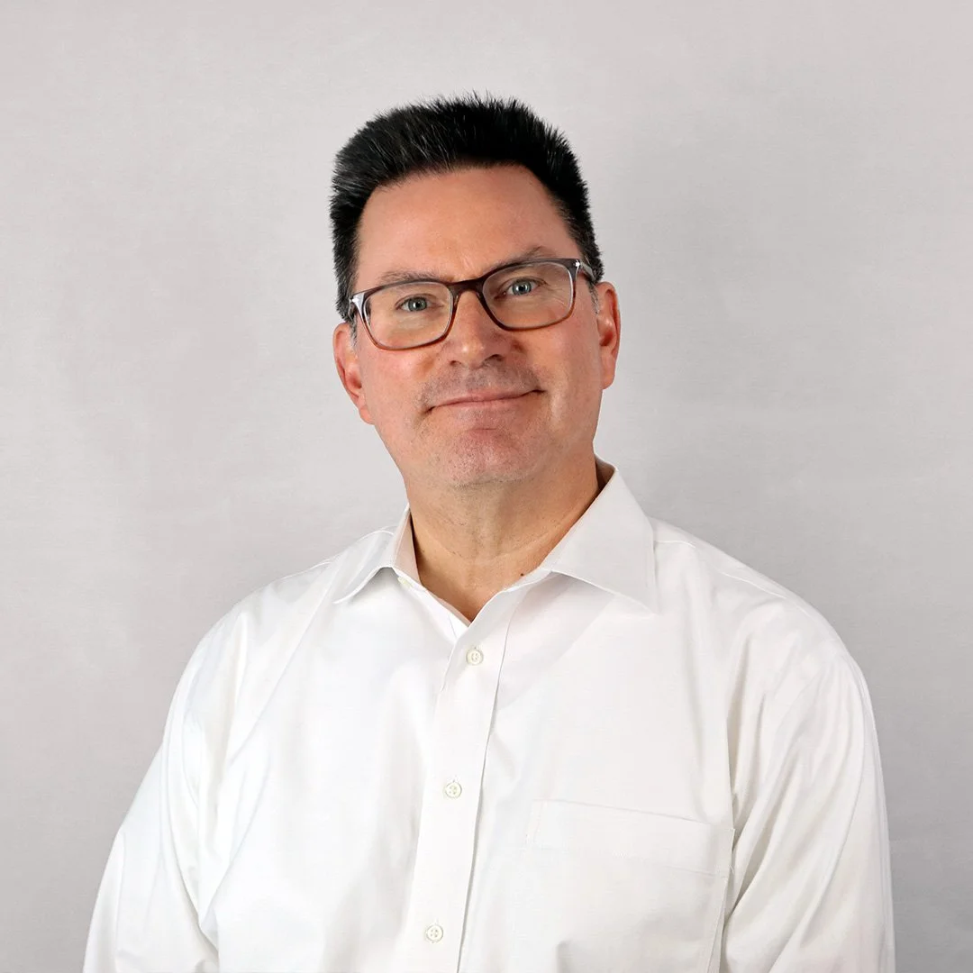 Portrait of a man with black hair, glasses, wearing a white dress shirt, smiling slightly, against a plain light background.