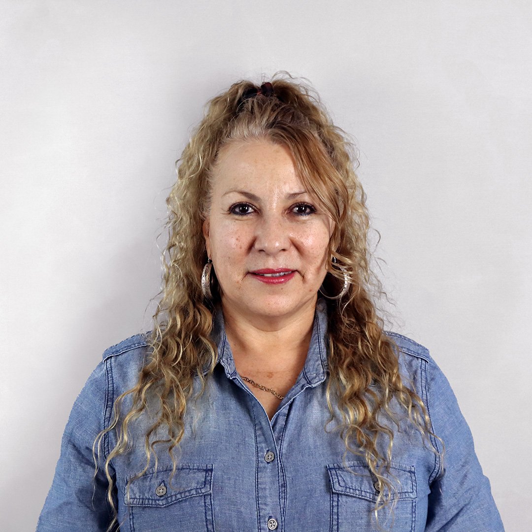 A woman with curly blonde hair wearing a denim shirt, posing against a plain white wall.
