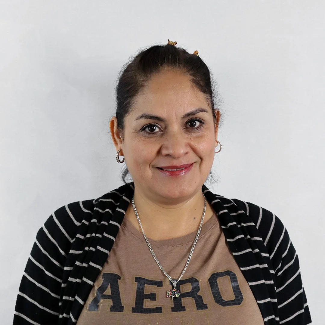 A woman smiling at the camera, wearing a beige Aero shirt, a black and white striped jacket, and silver jewelry, standing against a plain white background.