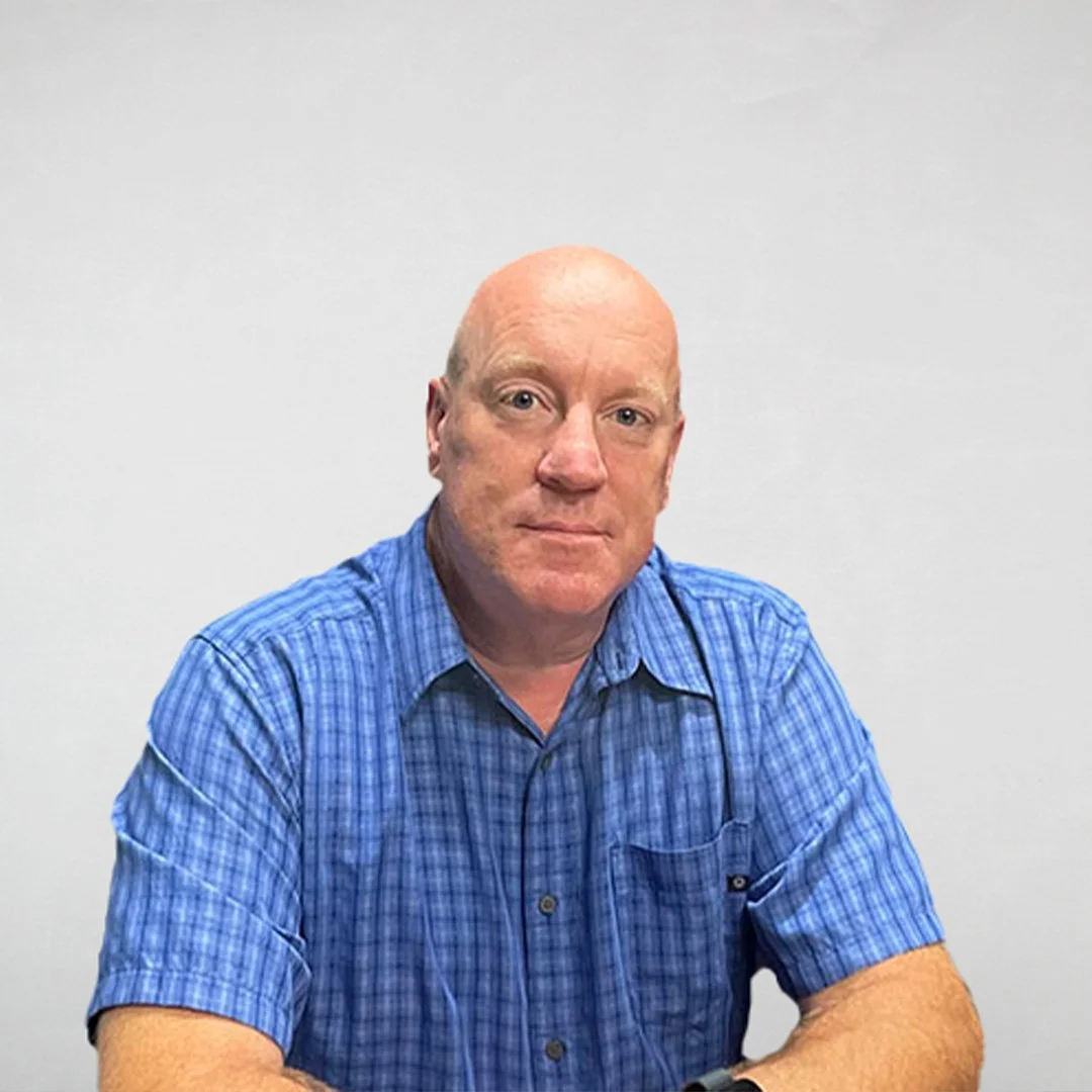 Man with a shaved head wearing a blue plaid shirt sitting at a table with a white wall background.