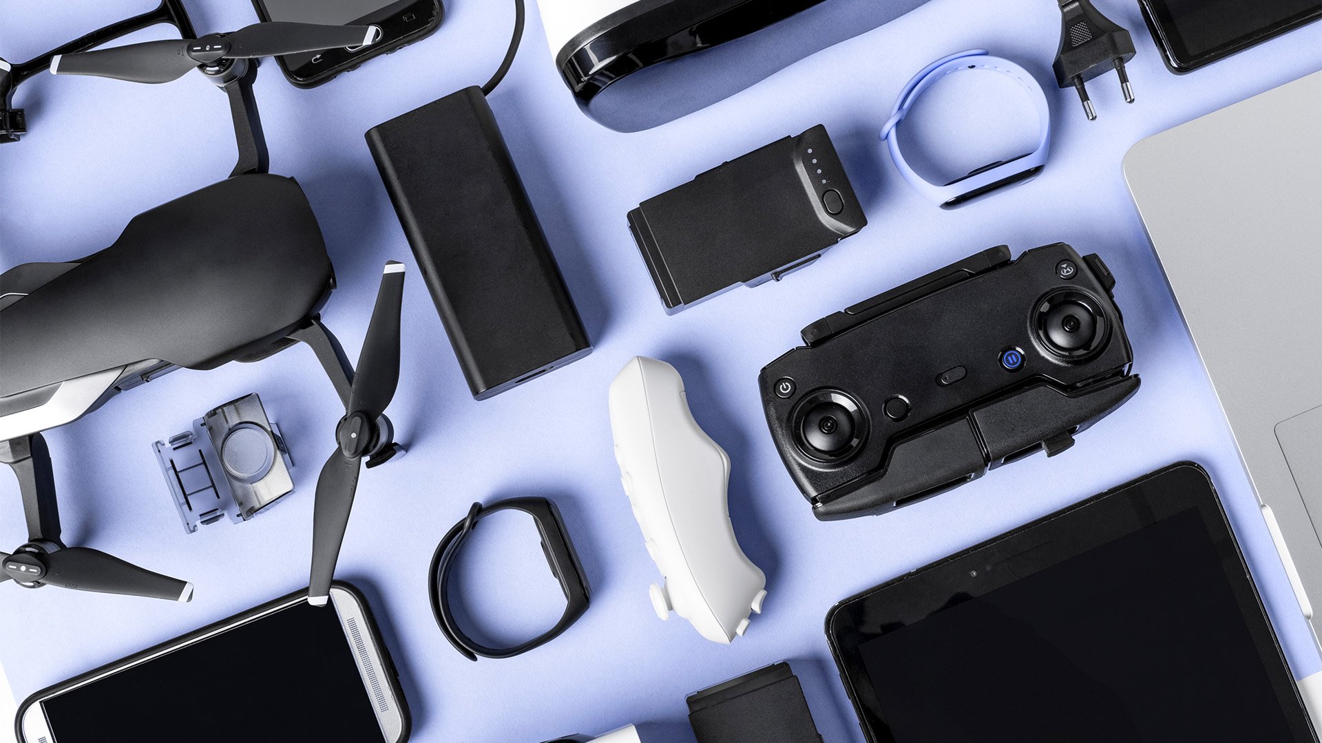 A collection of electronic gadgets on a light blue surface, including a drone with a camera, a remote control, headphones, a smartphone, a tablet, a laptop, a camera baffle, a battery, and various chargers.