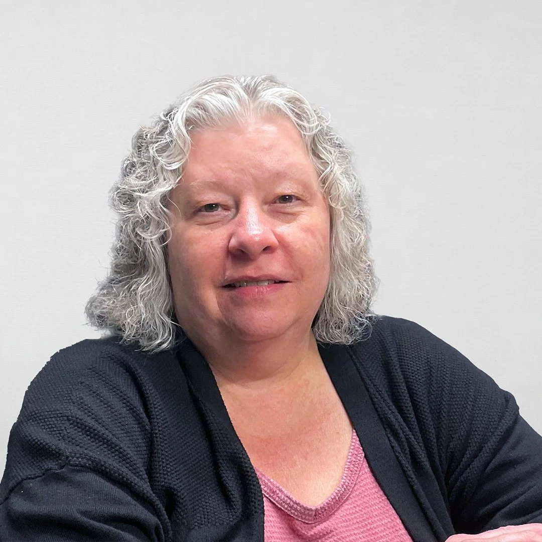 Portrait of a woman with curly gray hair wearing a black jacket and a pink shirt, smiling softly, against a plain light gray background.