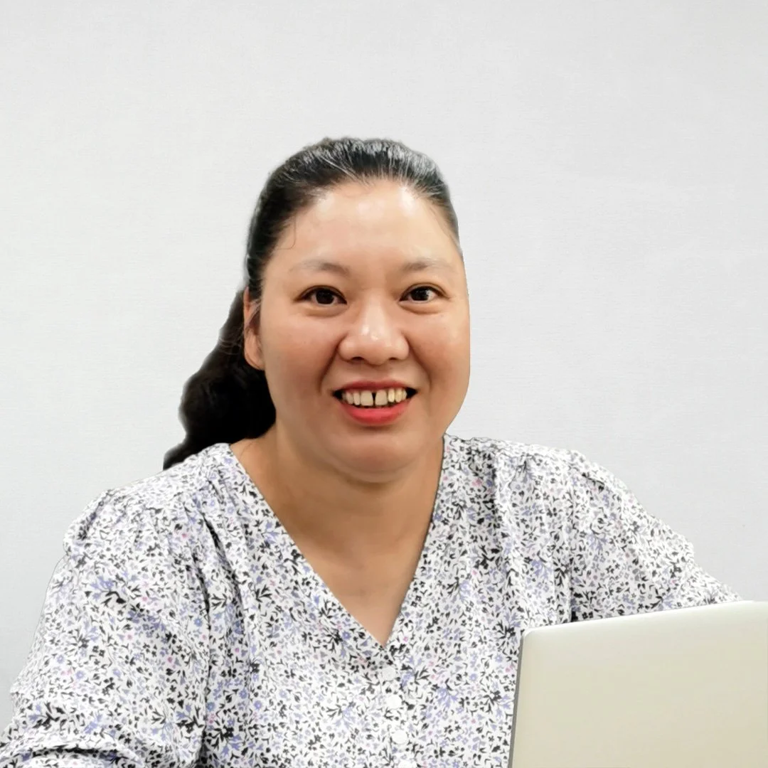A woman with dark hair in a ponytail, smiling, sitting at a desk with a white laptop, wearing a patterned blouse, against a plain white wall background.