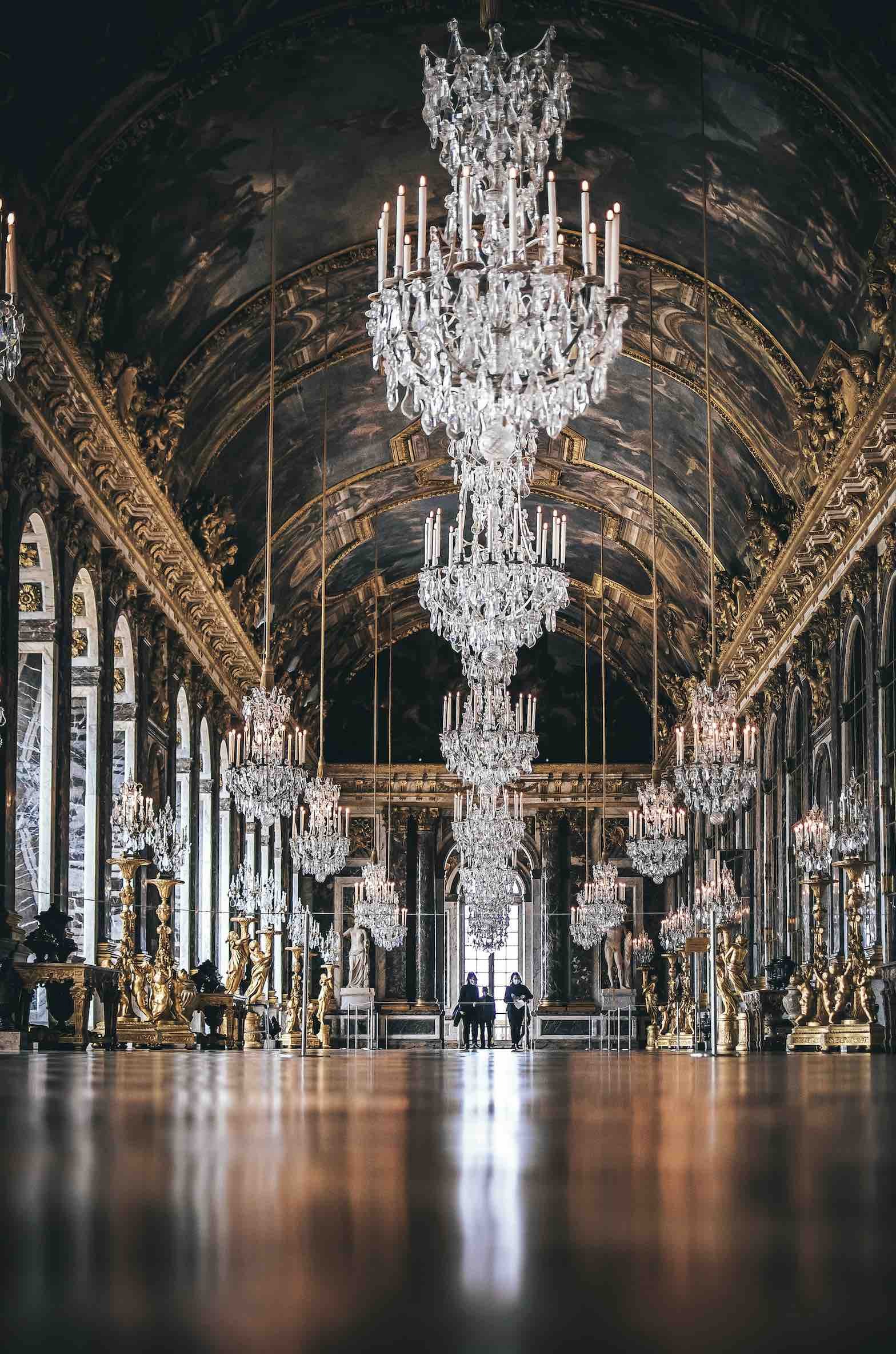 Luxurious interior hall with multiple large crystal chandeliers hanging from a high, elaborately decorated ceiling, featuring gold accents and tall windows with intricate frames.