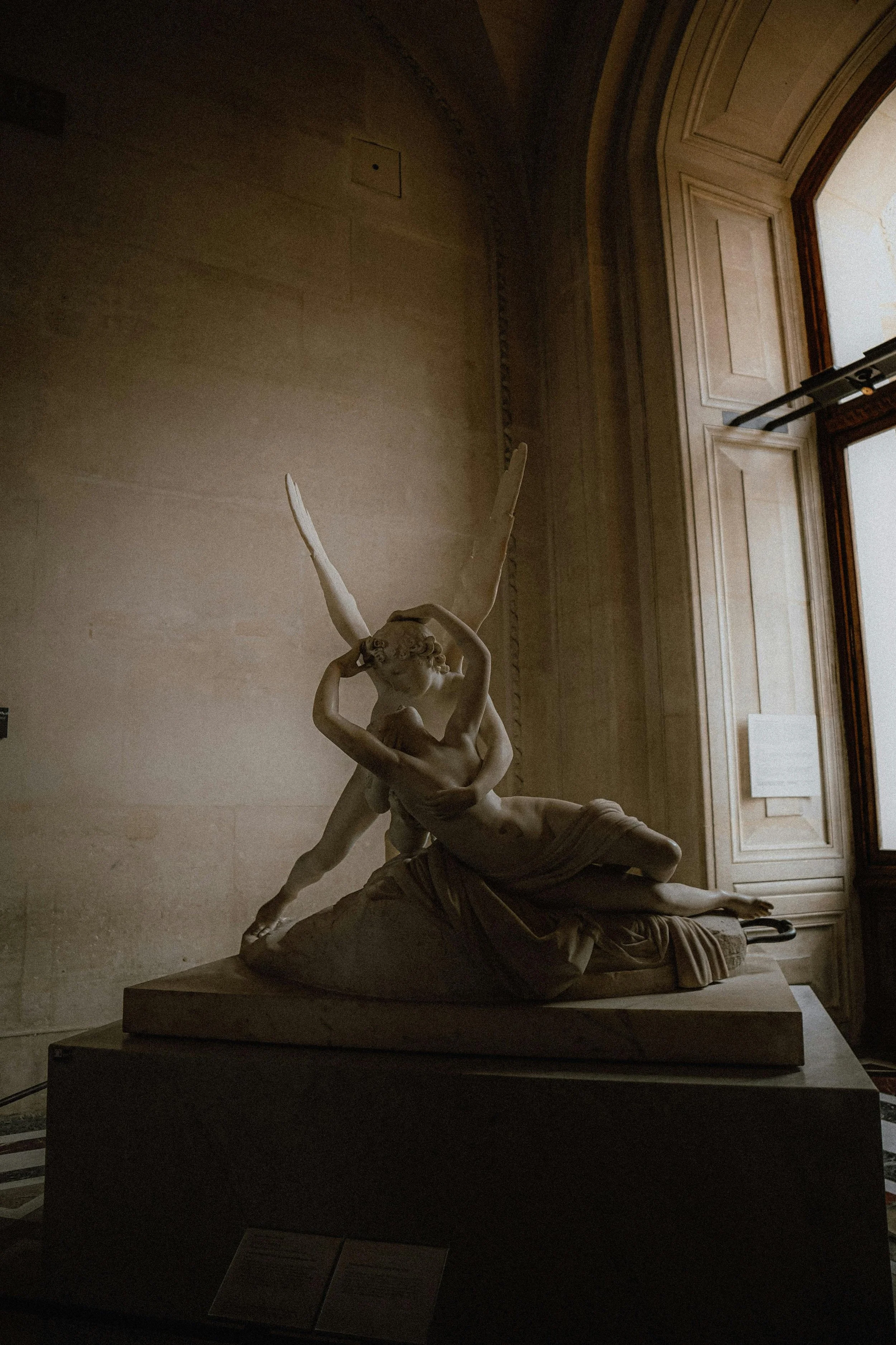 A classical marble sculpture of a woman with wings, reclining on a rock, with a male figure embracing her, displayed in a museum gallery with high windows and decorative molding.