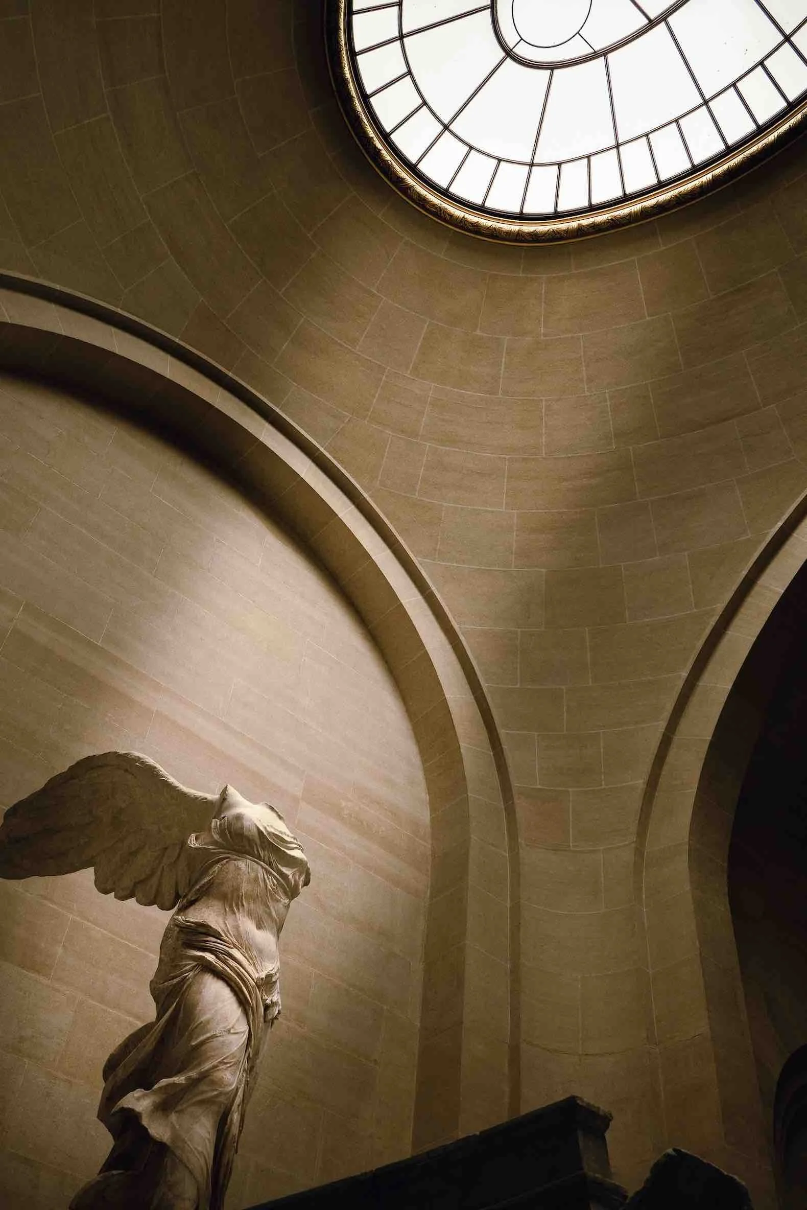 Interior view of a museum featuring a classical statue of a winged figure, with a large, circular, stained-glass skylight on the ceiling illuminating the scene.