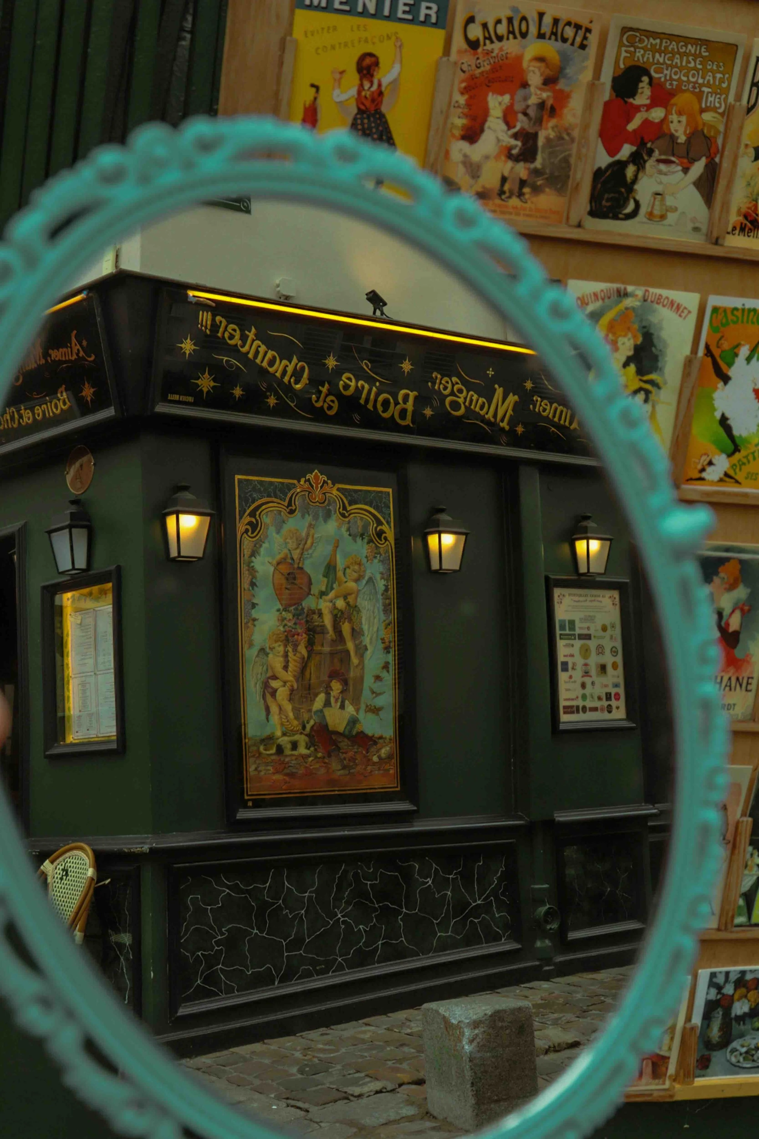 Reflection of a decorative green building with lanterns and a central painting, seen through an ornate teal mirror frame, with books displayed on shelves in the background.
