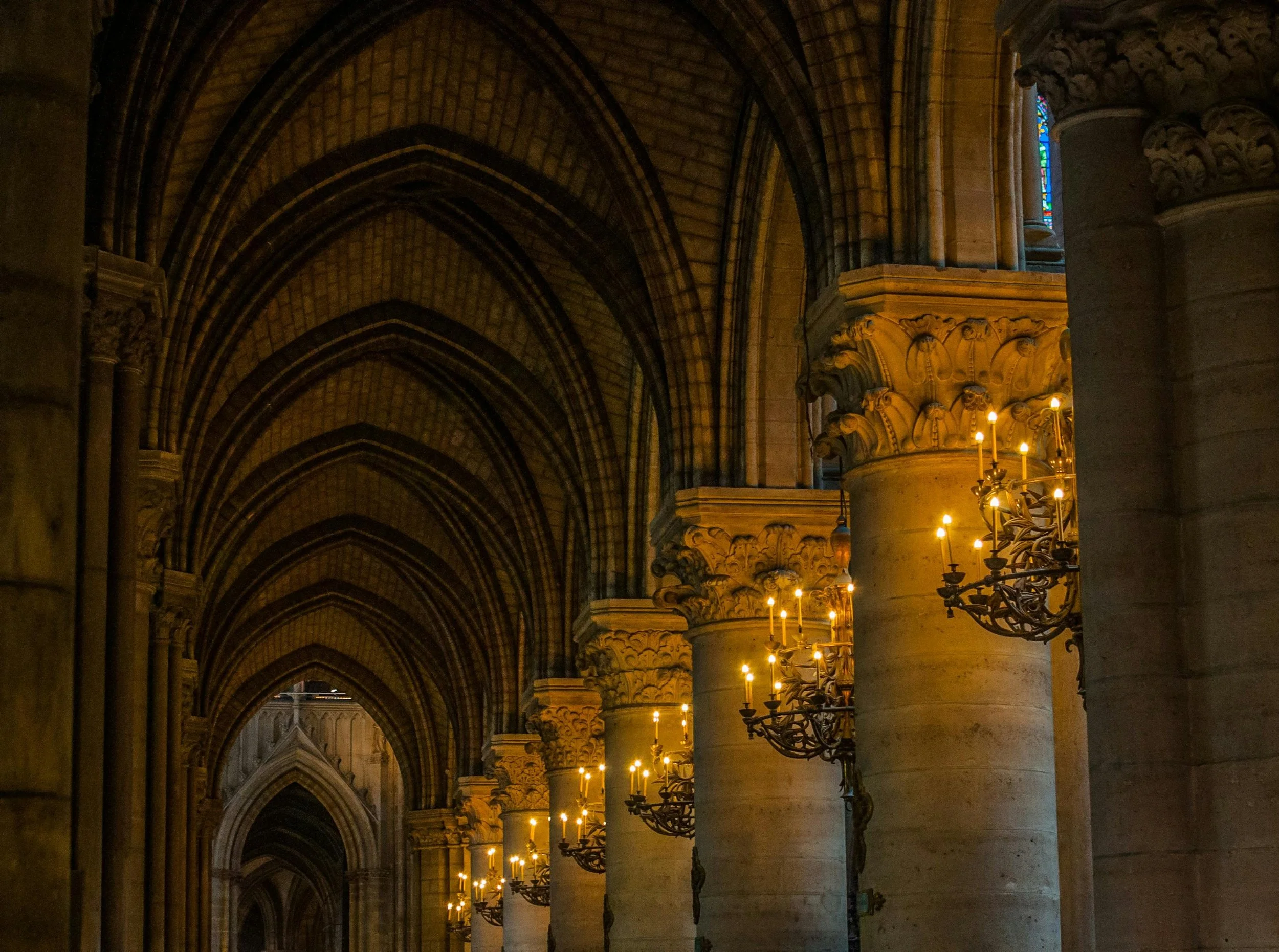 Interior of a cathedral with tall stone arches, ornate columns, and candle chandeliers along the corridor.