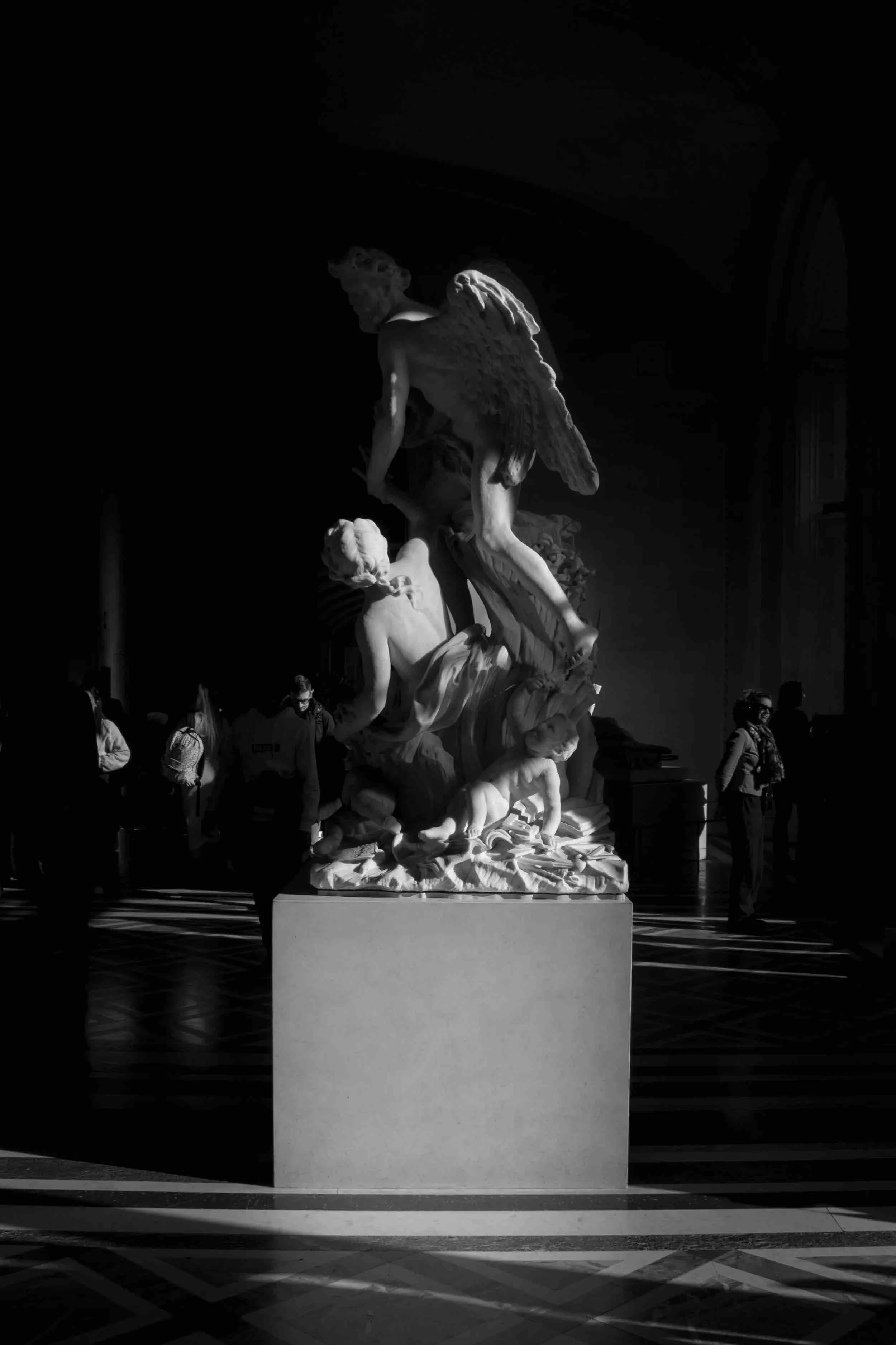 Black and white photo of a classical marble sculpture depicting angels and mythological figures, displayed in a museum with visitors in the background.
