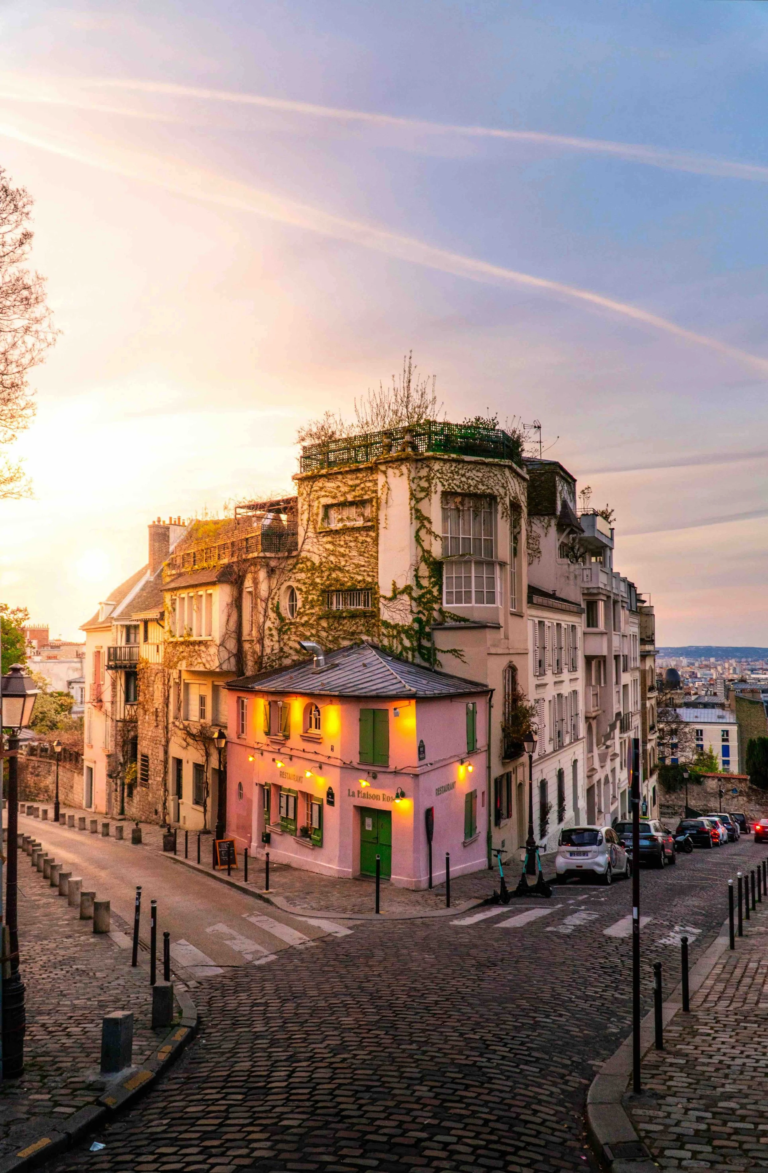 A charming pink building with green shutters and a restaurant sign, located at a curved cobblestone street in a European city, during sunset.