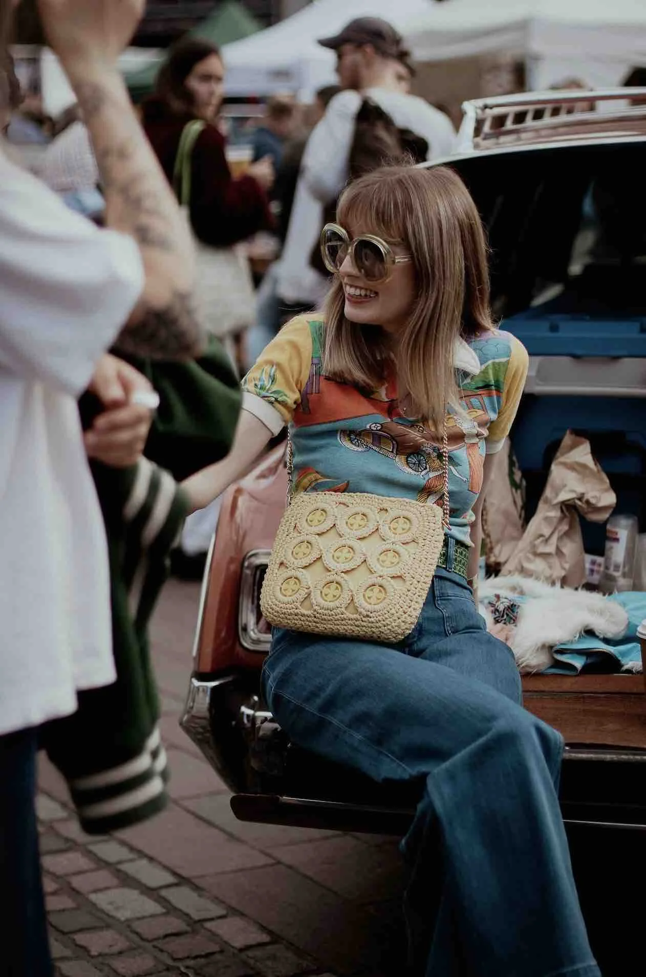 A woman with red hair wearing large sunglasses, a colorful patterned shirt, high-waisted jeans, and a yellow crocheted bag is smiling and chatting at an outdoor market or fair.