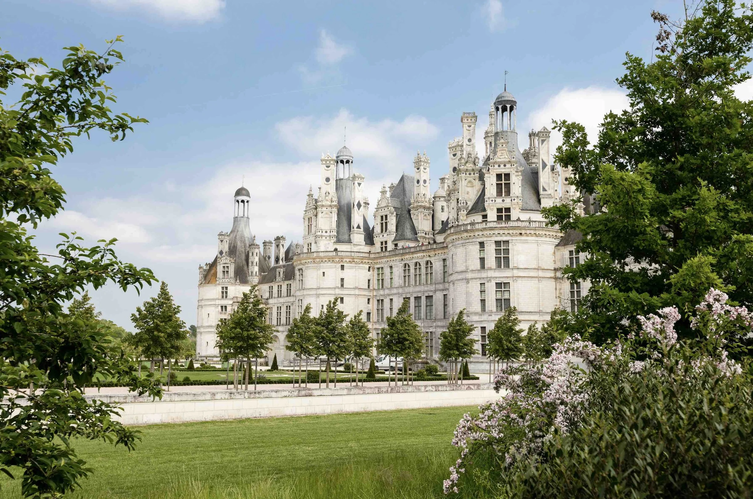 A large, ornate castle with numerous turrets and towers, surrounded by green trees and a well-manicured lawn, under a partly cloudy sky.