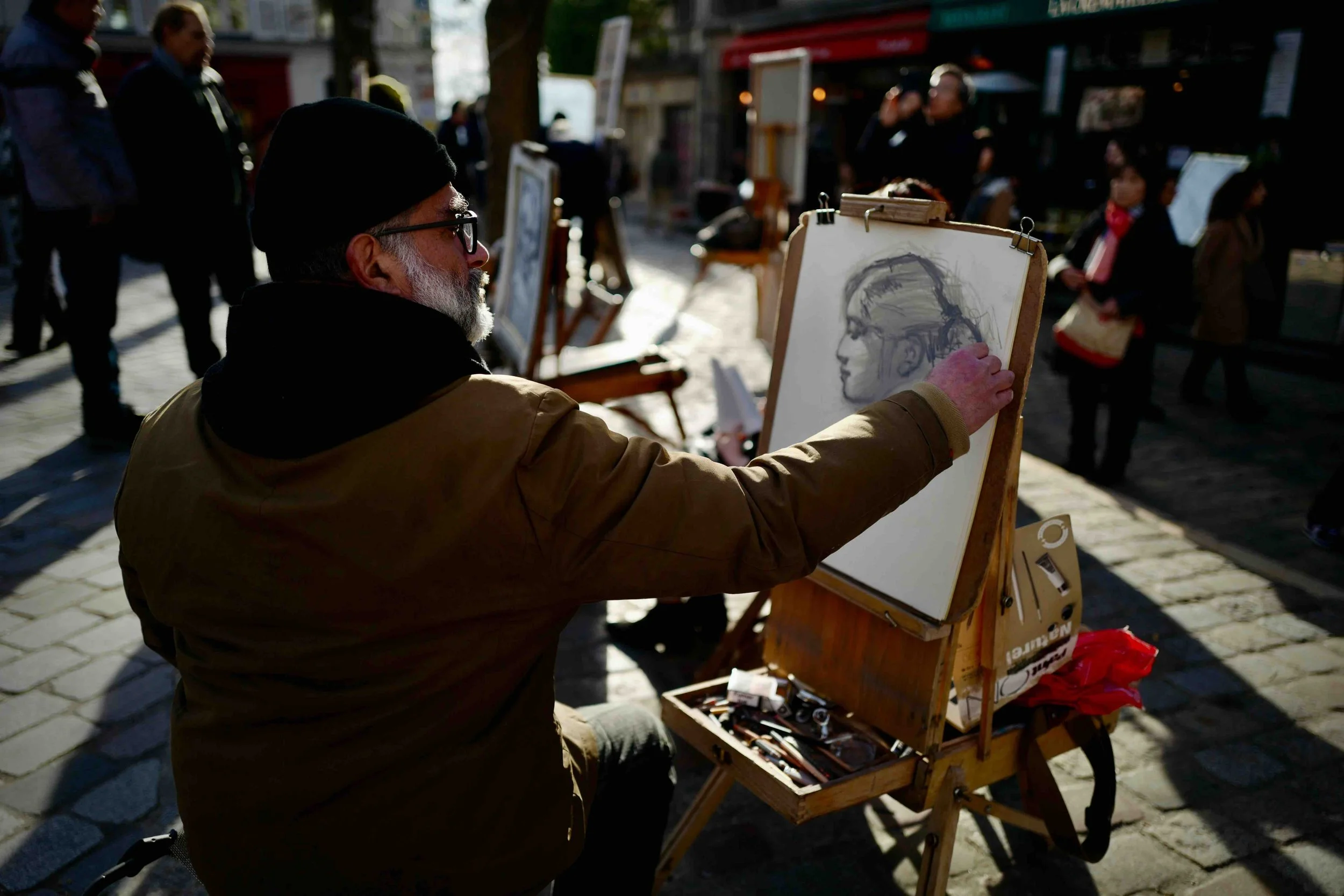A man with a beard, glasses, black beanie, and brown jacket is drawing a portrait of a woman on a sketchpad at an outdoor street market. Other people are walking and browsing nearby.