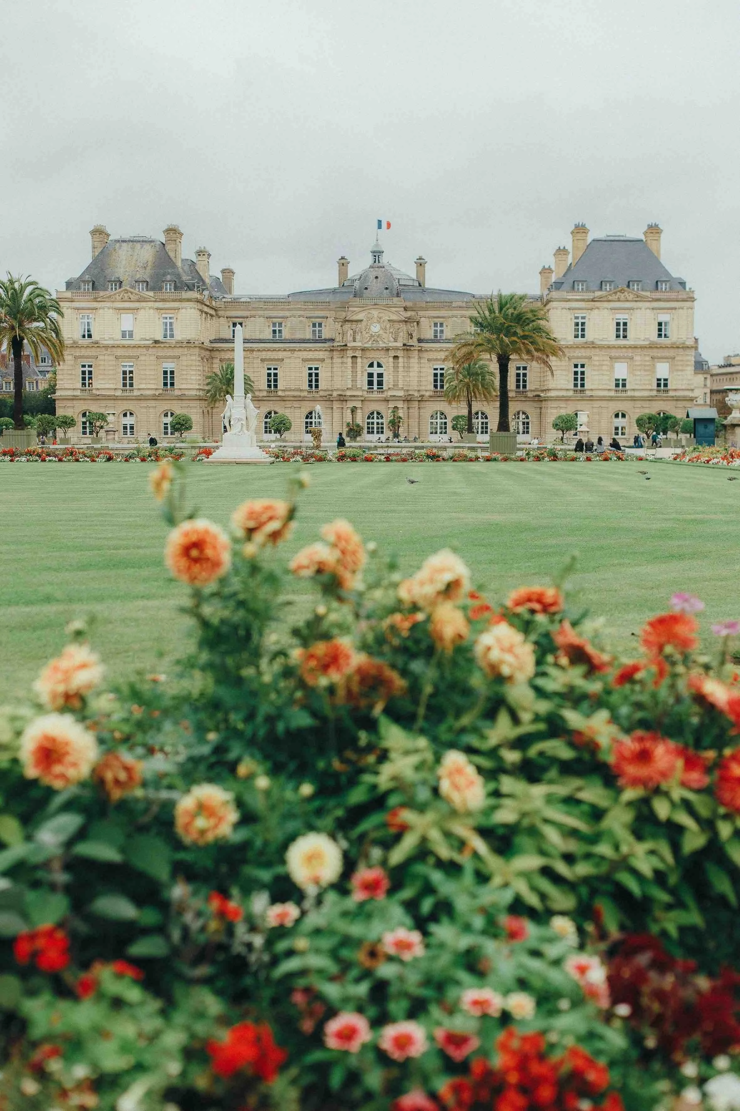 A large historic building with a European architectural style, surrounded by a lawn and colorful flowers, with palm trees and a statue in front.