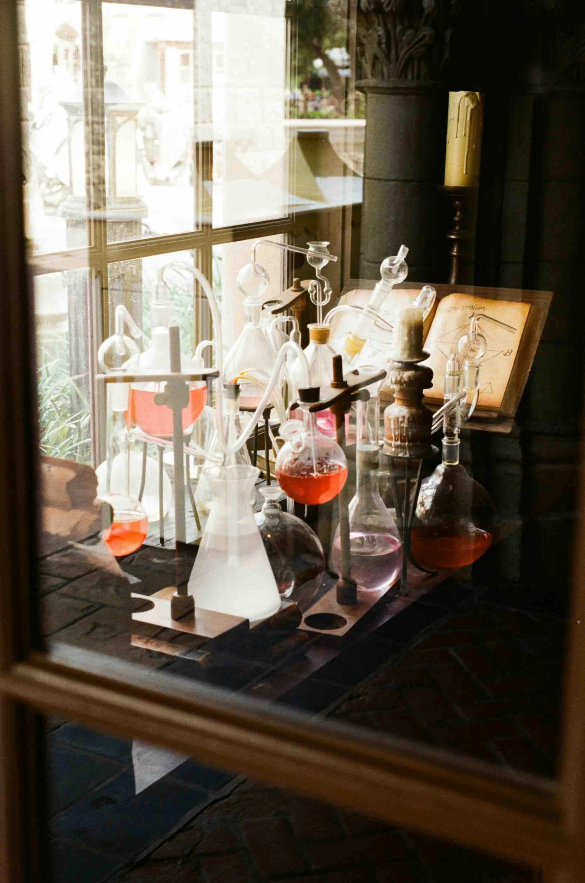 Science laboratory setup with glass flasks containing red and clear liquids, scientific equipment, and an open book with diagrams on a wooden table near a window.