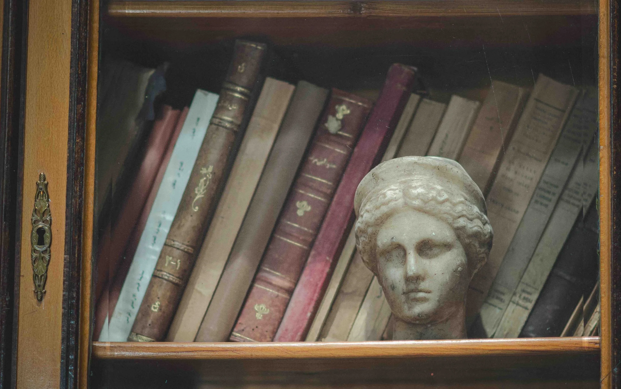 A bookshelf with old books and a small sculpture of a woman's head inside a glass cabinet.