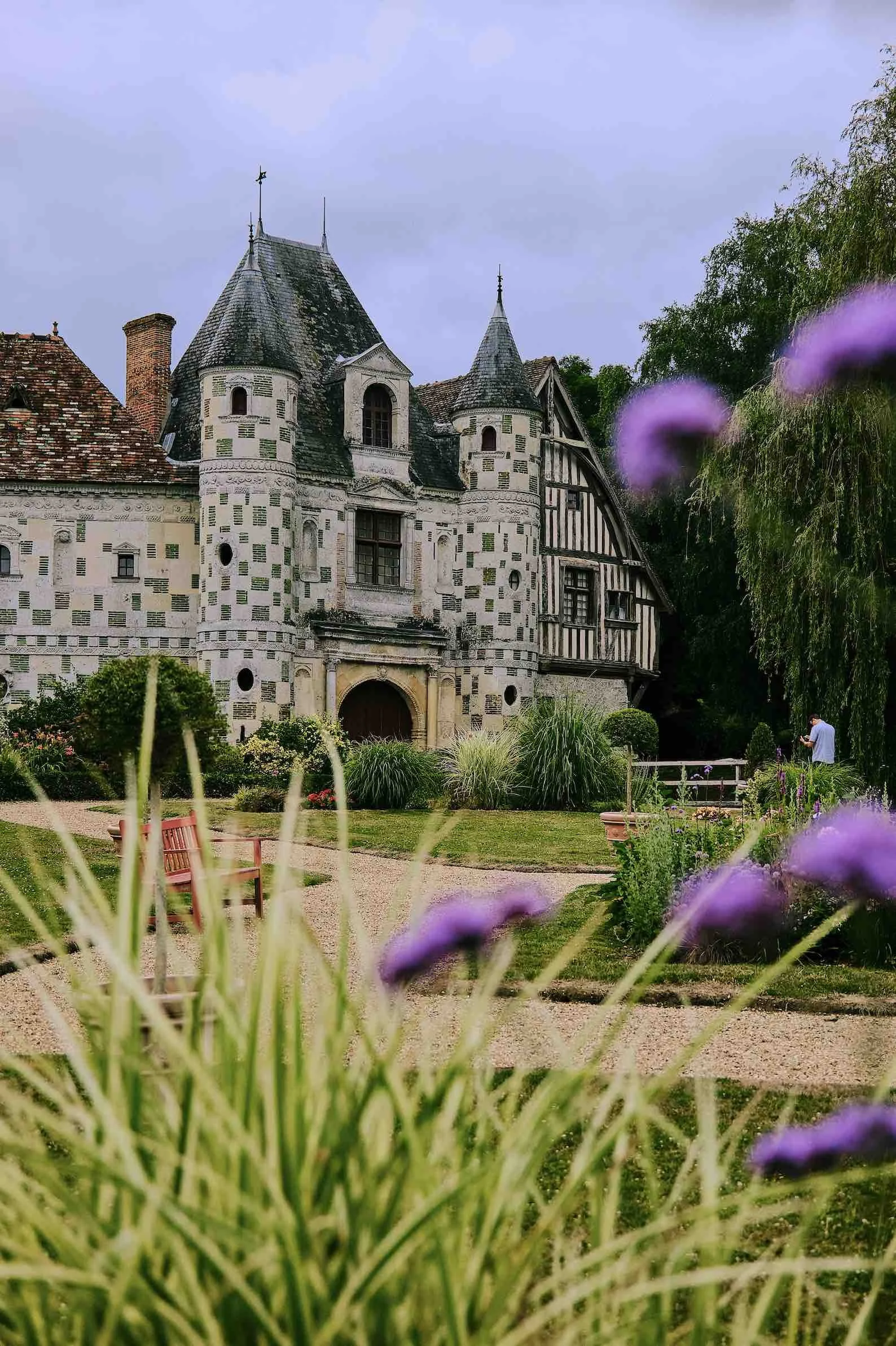 An old castle with stone walls and turrets, surrounded by a garden with purple flowers, bushes, and a bench.