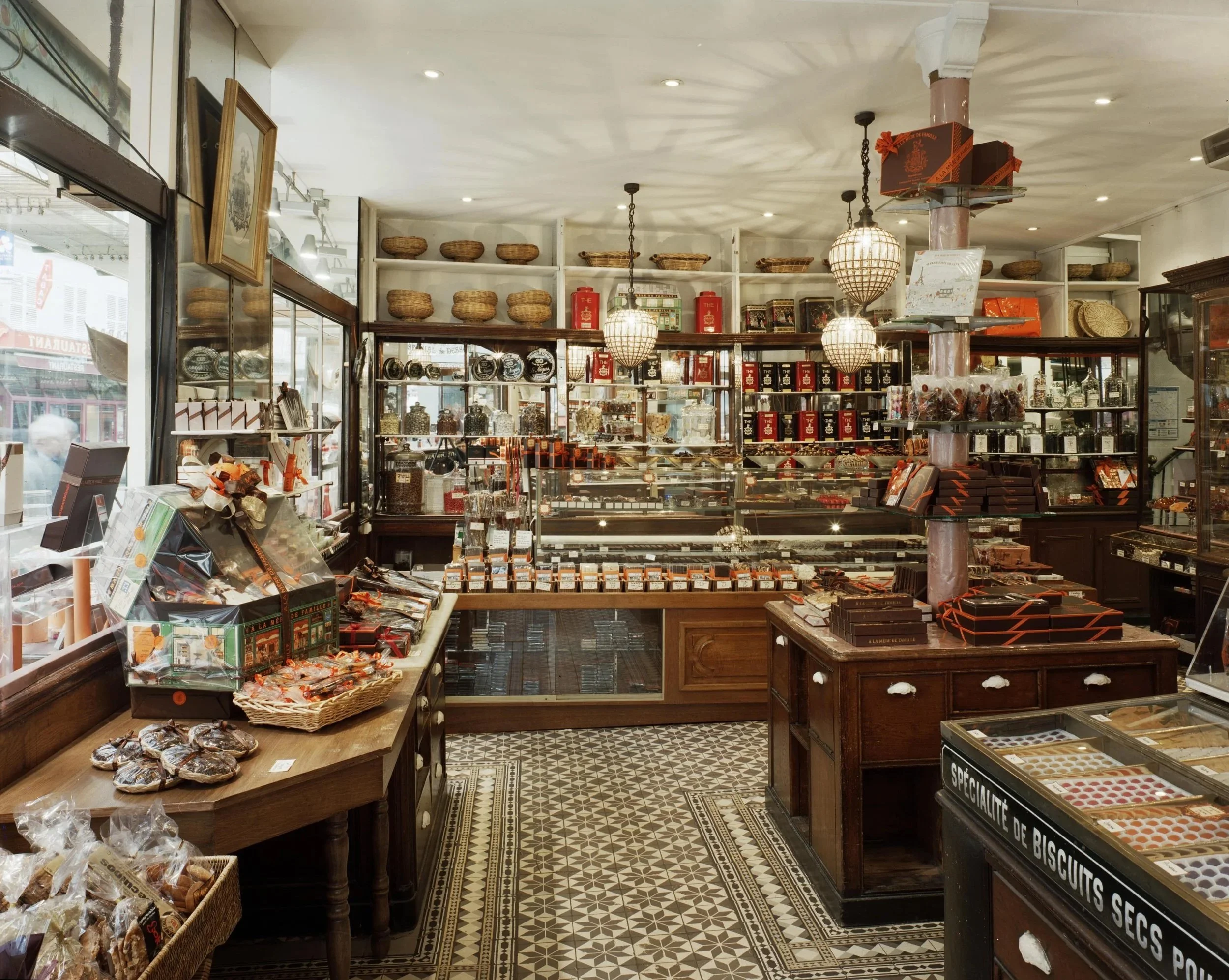 Interior of a traditional European-style chocolate shop with wooden furniture, glass display cases filled with chocolates, and shelves with tins and baskets, illuminated by hanging pendant lights.