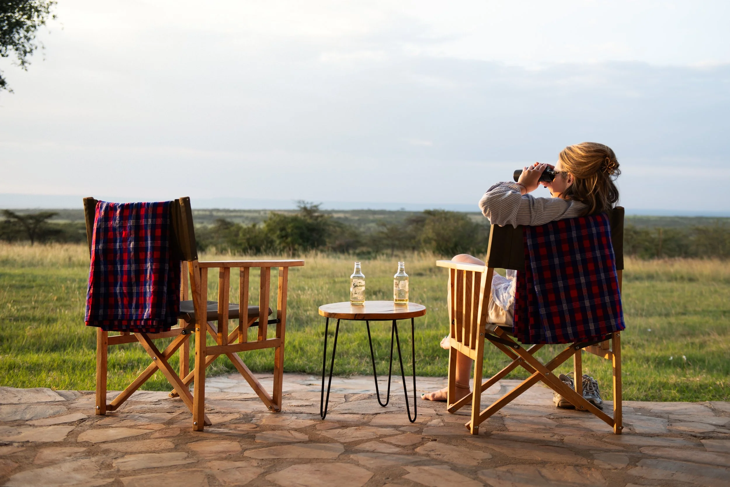 A woman sitting on a wooden chair on a stone patio, looking through binoculars into a grassy landscape with scattered trees, two glass bottles on a small round table, and another empty wooden chair beside her.