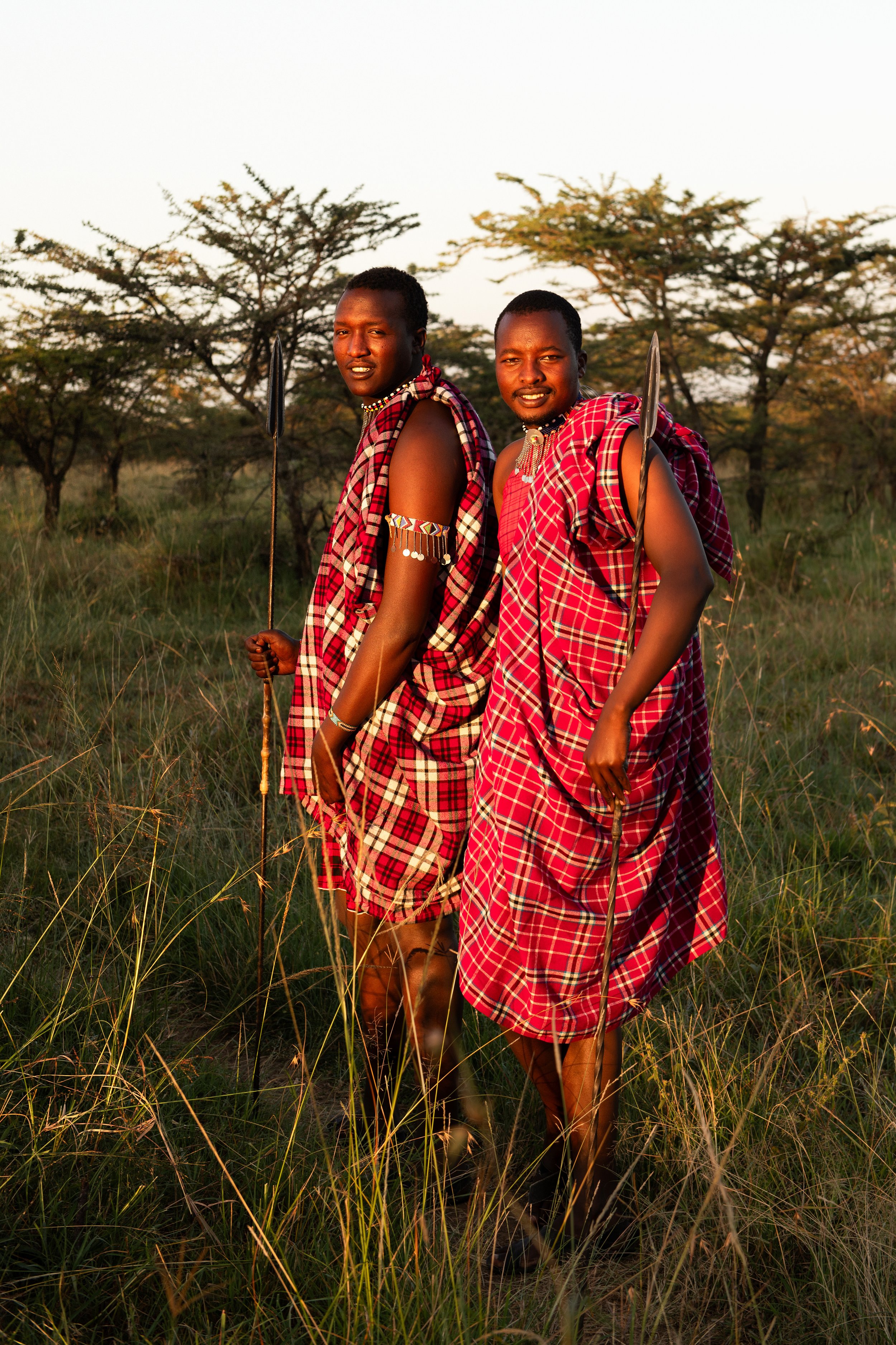 Two Maasai men dressed in traditional red and black plaid shukas, standing in a grassy savannah with trees in the background at sunset, holding spears.