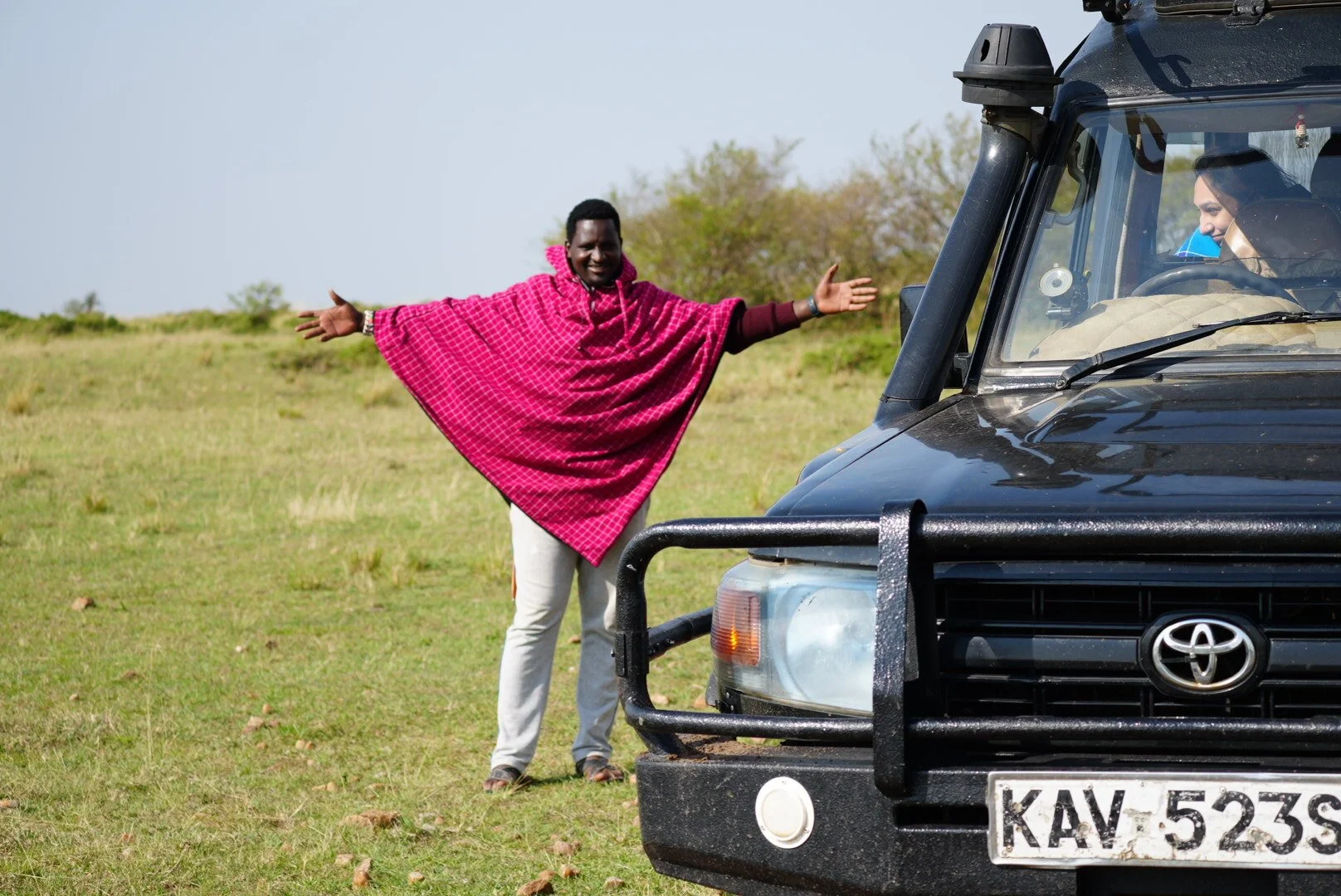 A man standing outside in a grassy field, smiling and spreading his arms, wearing a red checkered shawl. A black Toyota vehicle with a license plate KAV 523S is parked nearby, with a person visible inside the vehicle.