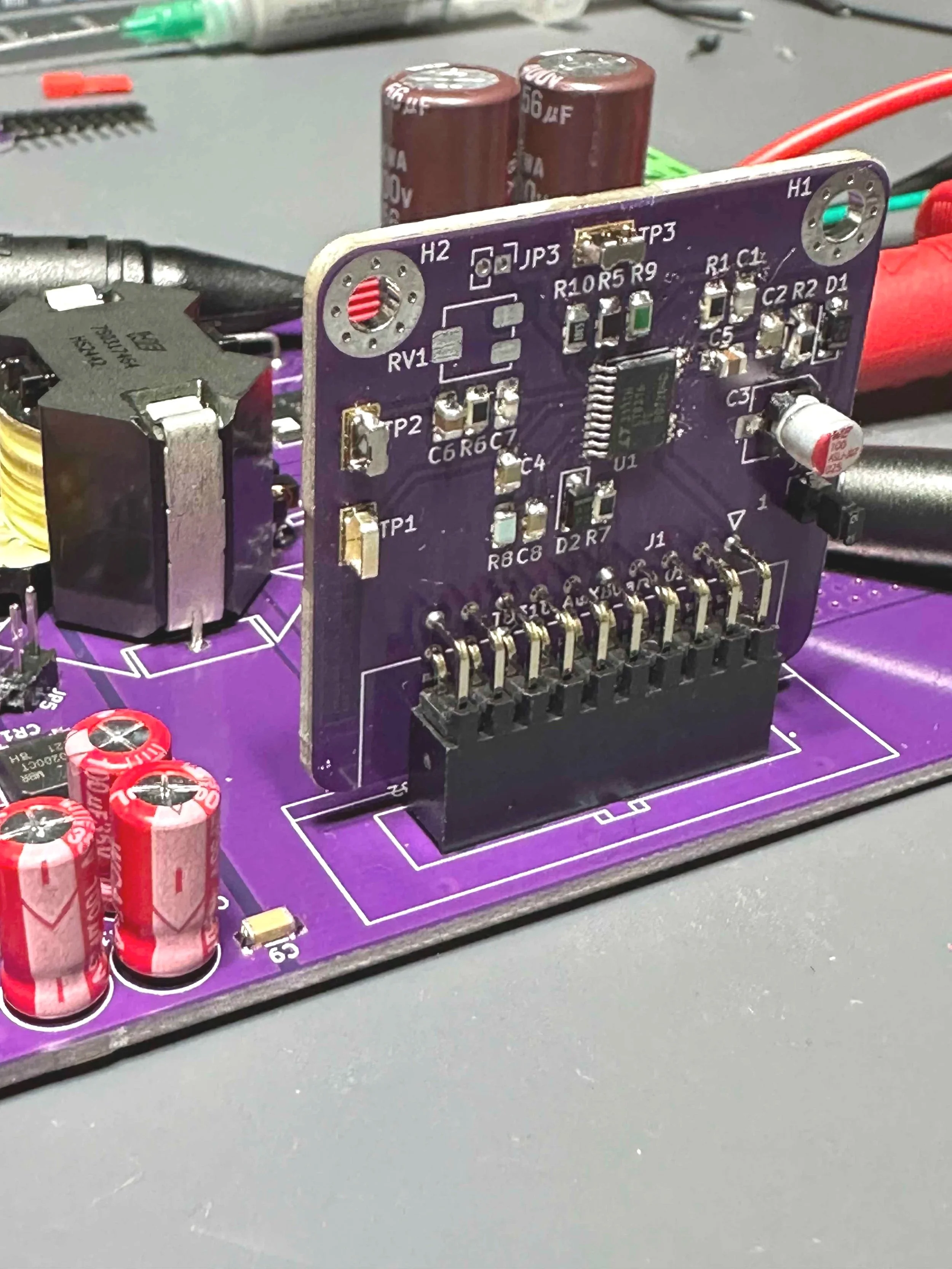 Close-up of a purple printed circuit board with electronic components, including capacitors, resistors, and an integrated circuit, mounted on a gray work surface.