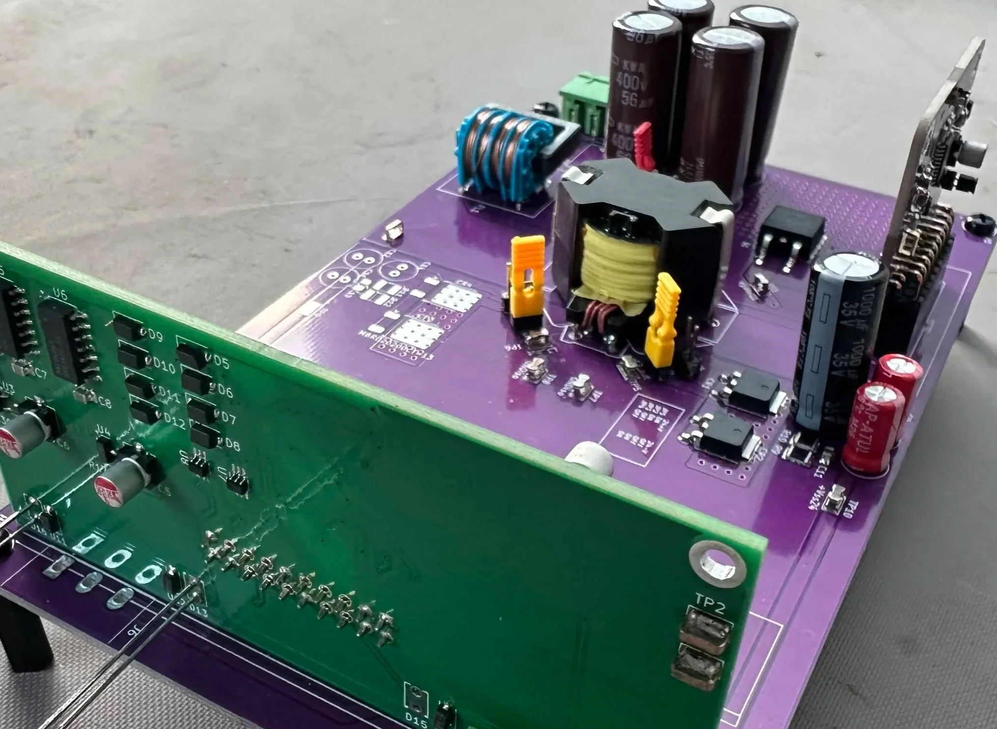 Close-up of an electronic circuit board with various components including capacitors, resistors, and inductors, mounted on a purple and green printed circuit board with electronic connections and labels.