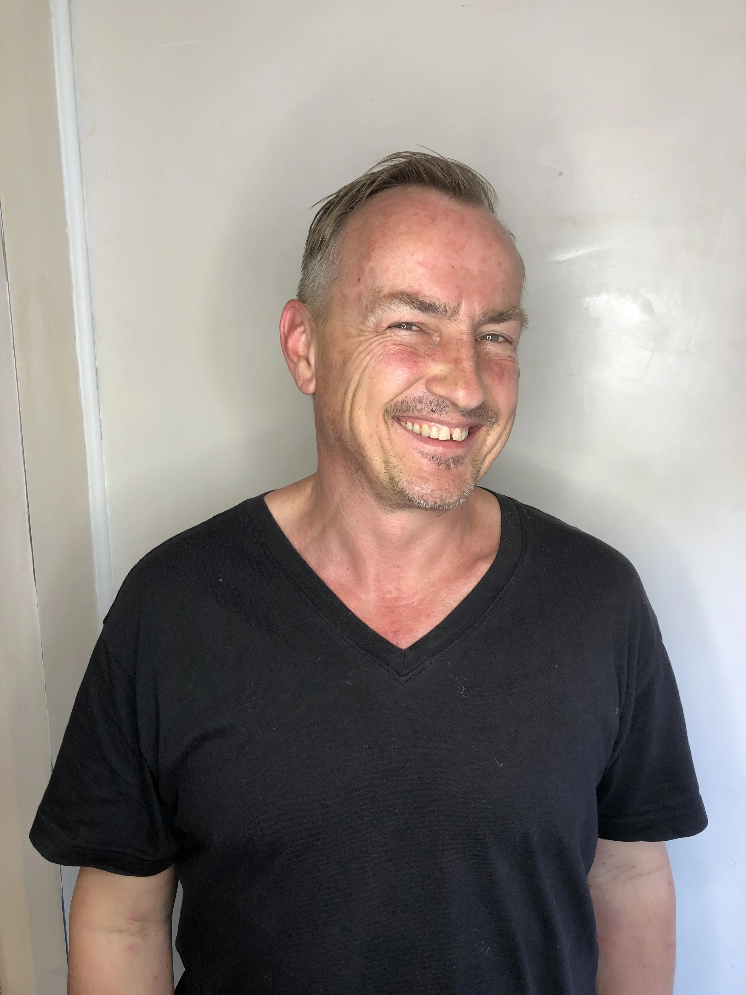 A smiling man wearing a black V-neck T-shirt standing in front of a plain white wall.