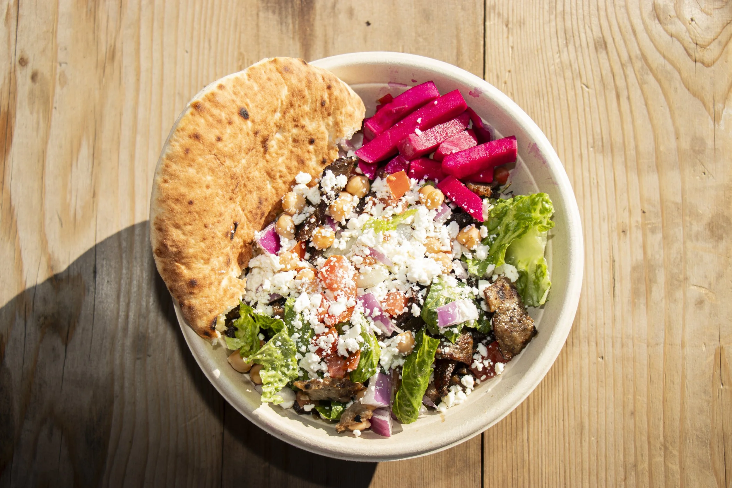 A bowl of salad with lettuce, tomatoes, red onions, chickpeas, crumbled cheese, and pieces of grilled meat, topped with pink pickled vegetables and served with a piece of toasted flatbread on a wooden table.