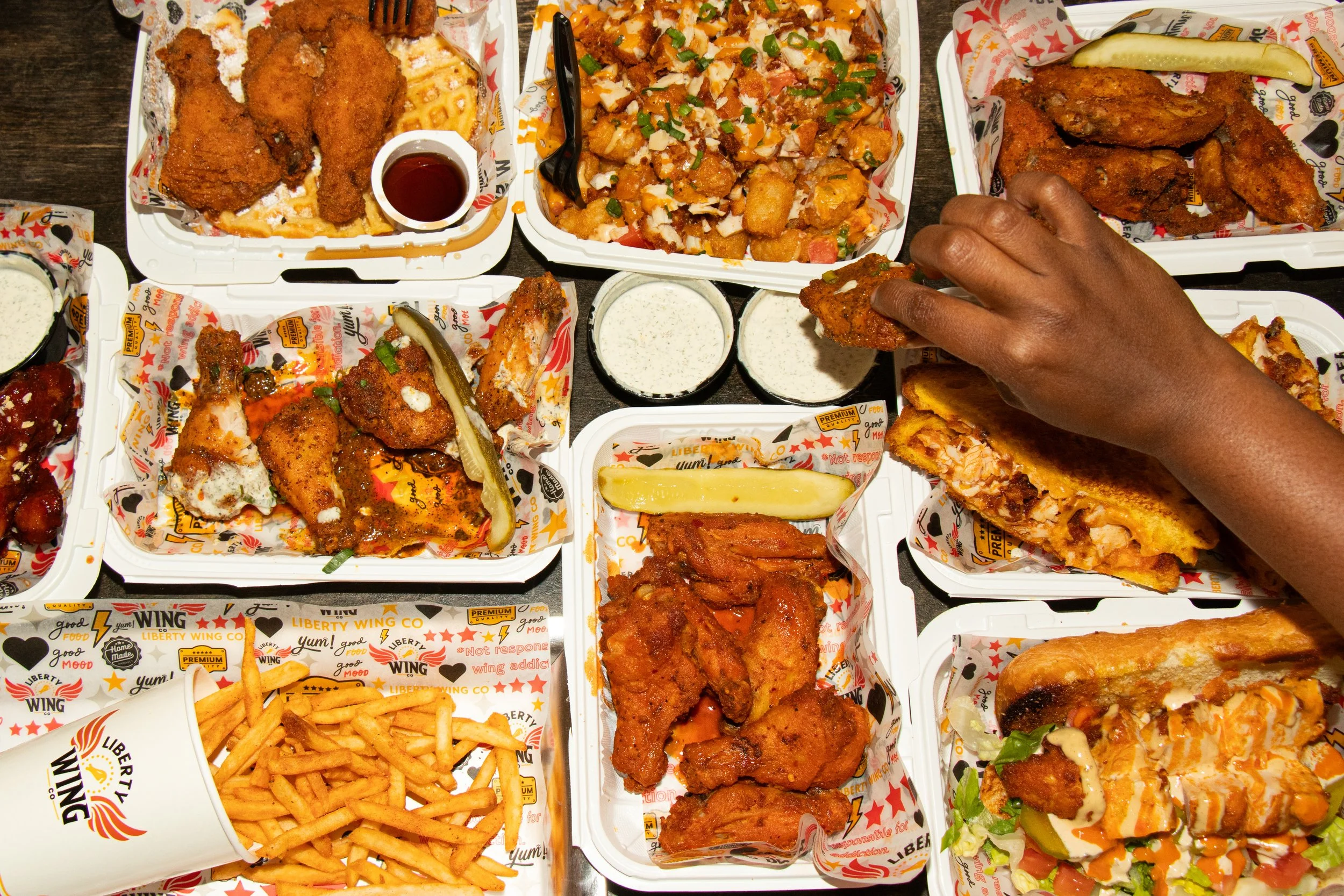 A top-down view of a large spread of fast food items in white and orange branded containers, including fried chicken, chicken wings, French fries, salads, and various chicken sandwiches, with a person's hand reaching for one of the sandwiches.