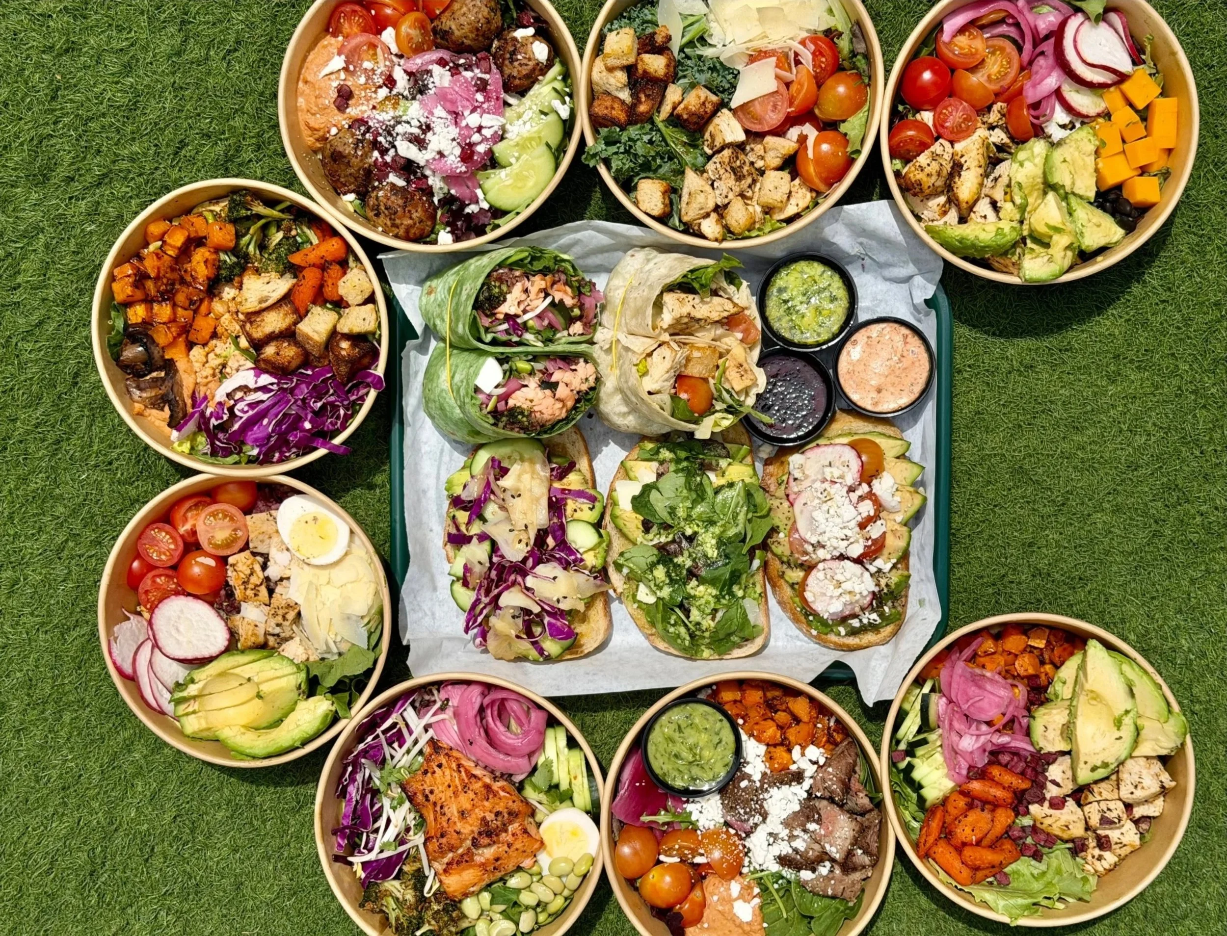 Multiple bowls of salads and wraps arranged on grass, containing various vegetables, meats, and toppings.