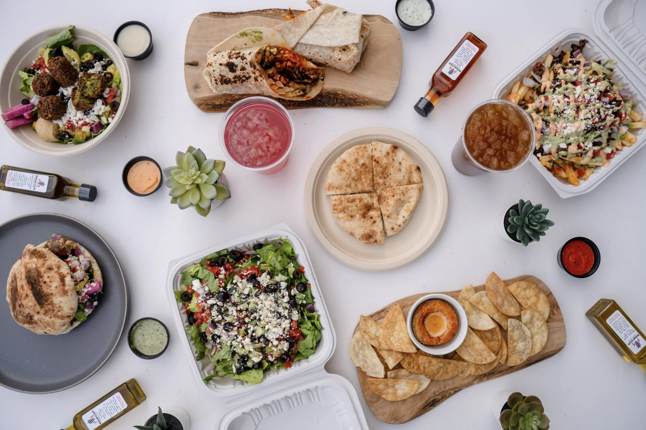 A spread of assorted Mexican dishes on a white table, including nachos, tacos, a burrito, salad, and beverages, with small potted succulents and condiments.