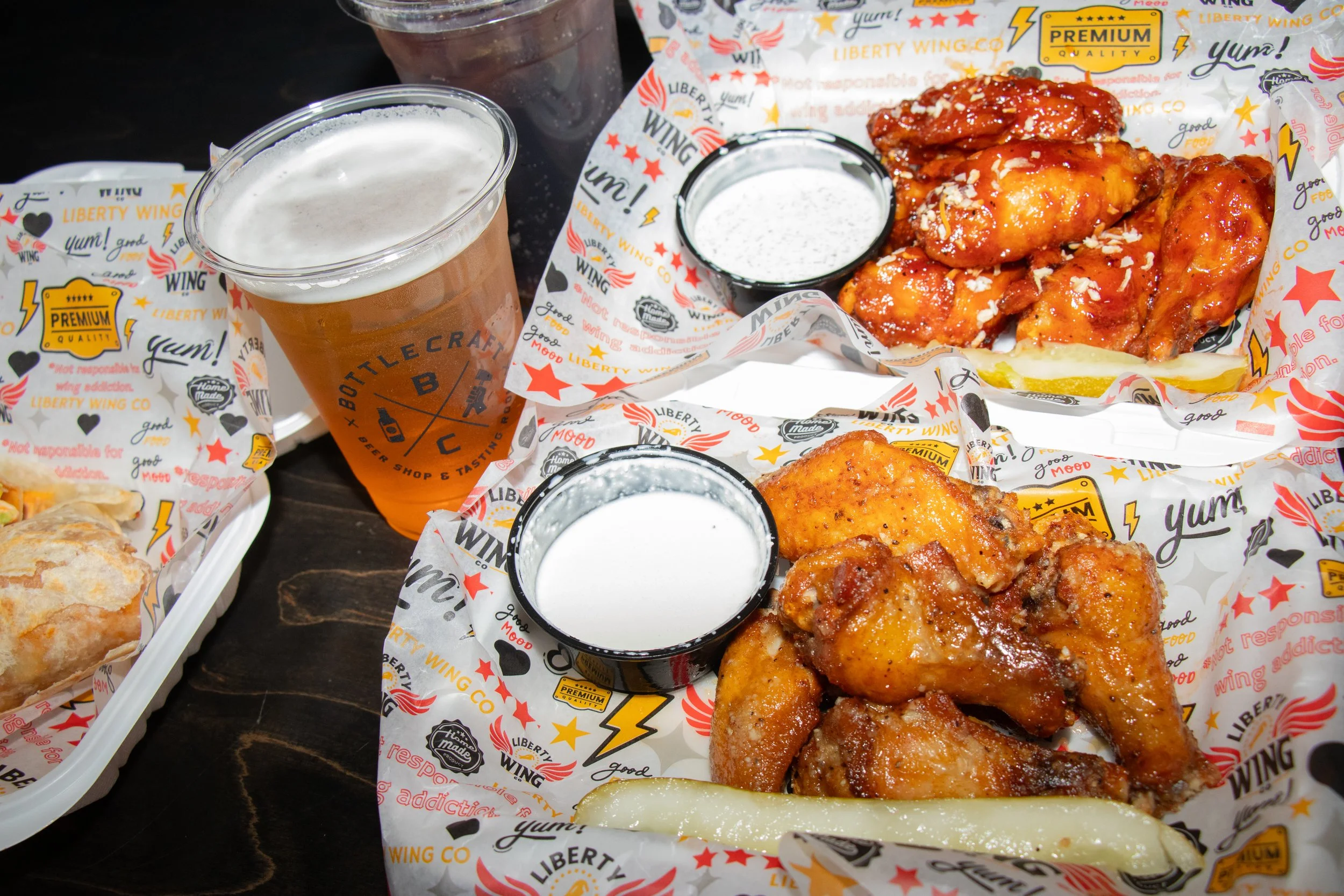 A tray of fried chicken pieces with dipping sauces, two small cups with white sauce, a glass of beer, and another fried item, all on branded paper from Liberty Wing.