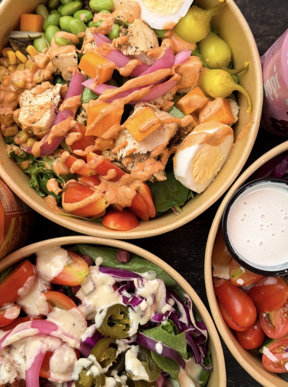 Assorted salads in bowls, including a salad with cherry tomatoes, shredded vegetables, and a creamy dressing, and a salad with leafy greens, sliced vegetables, and a light dressing, with a glass of milk and bottled drinks nearby.