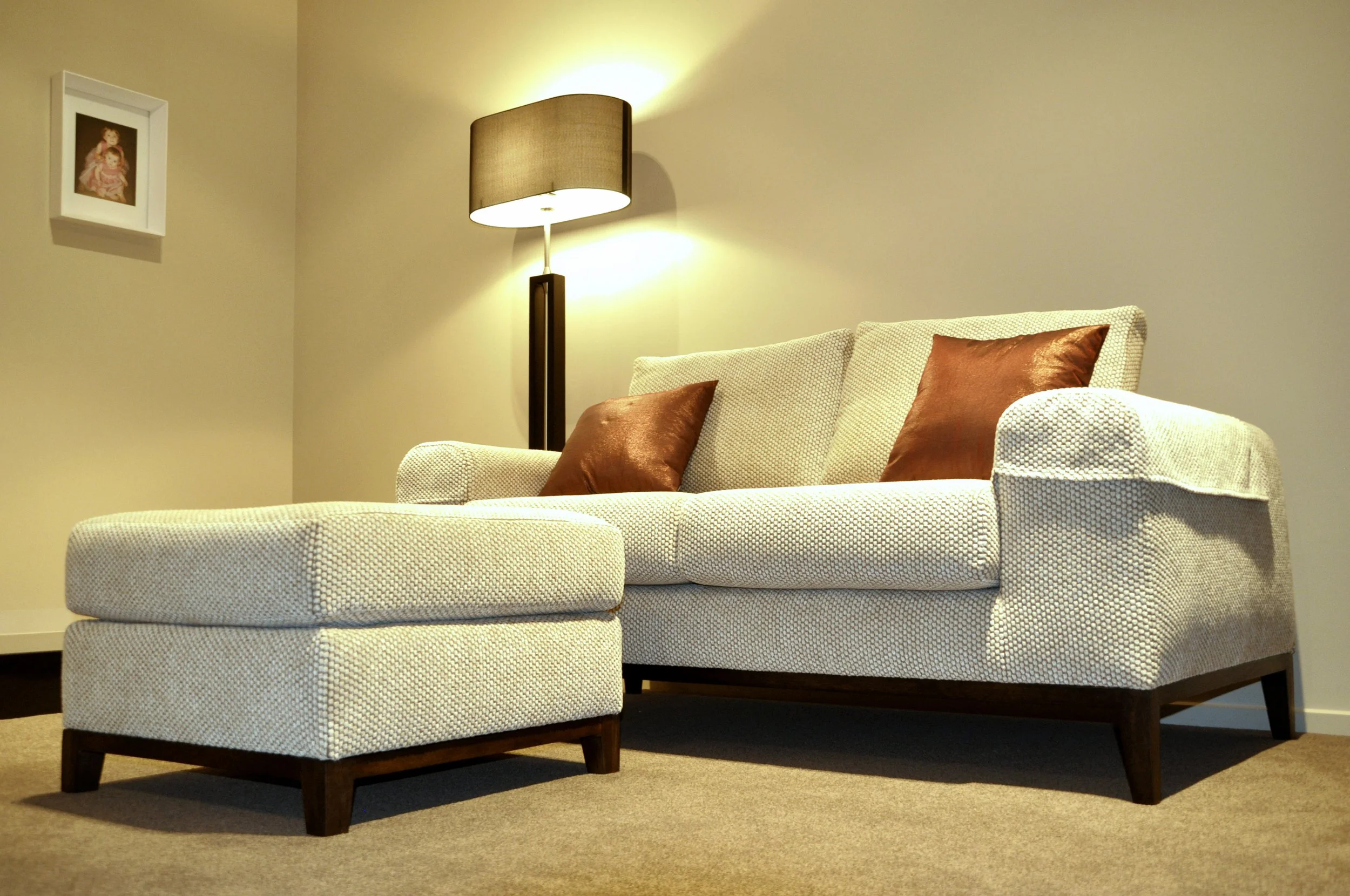 Living room with a beige sofa, matching ottoman, two brown throw pillows, a floor lamp, and a framed picture on the beige wall.