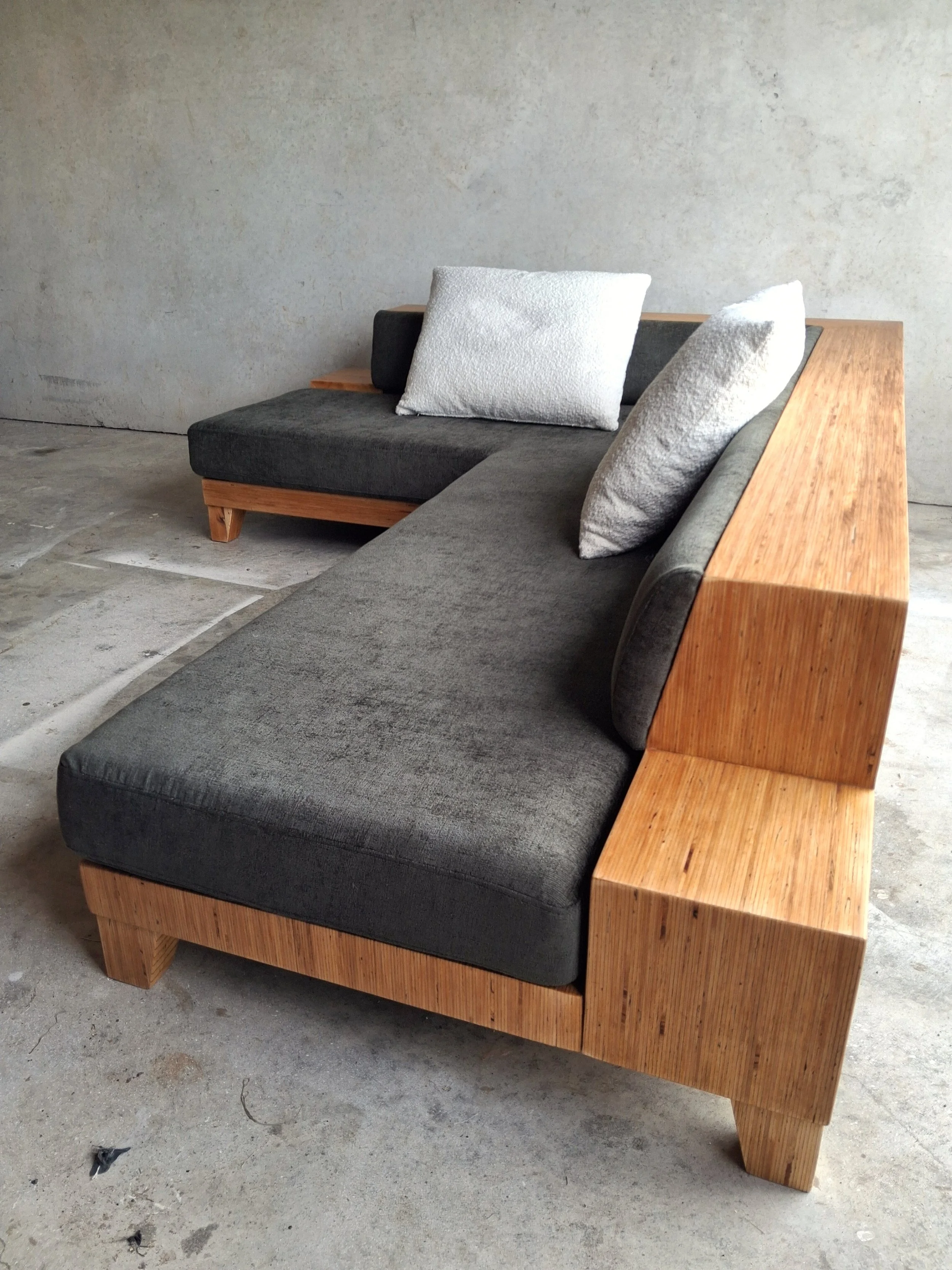 A modern sectional sofa with dark gray cushions and three light-colored throw pillows. The frame and armrest are made of natural wood, with angled wooden legs. The sofa is placed on a concrete floor against a plain, textured wall.