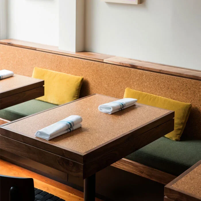 A restaurant table with two white napkins and rolled silverware, surrounded by beige and green booth seating with yellow cushions, in a modern dining area.
