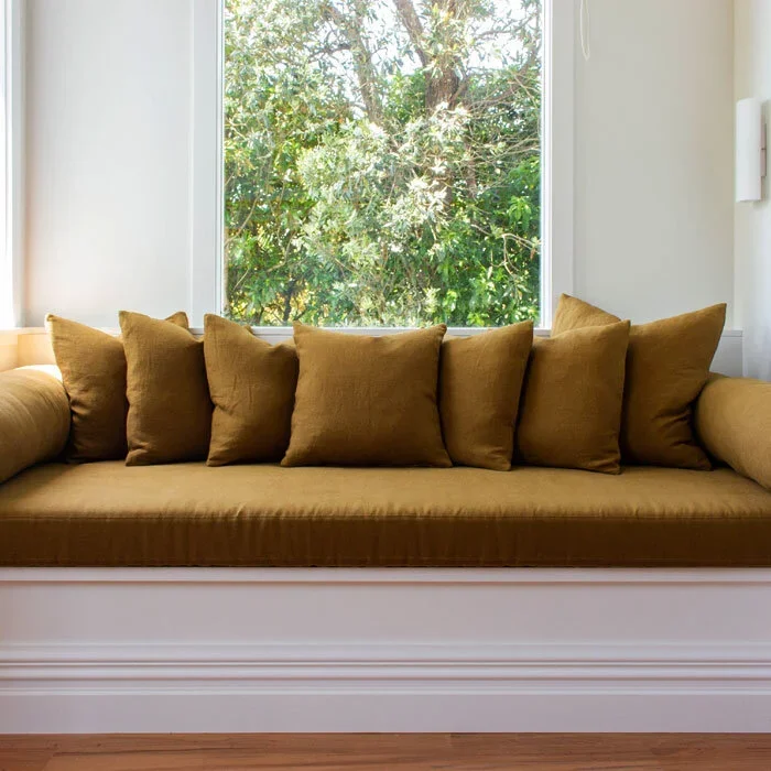 A window seat with a brown cushioned bench and six matching throw pillows, nestled in a brightly lit room with a large window showing green foliage outside.