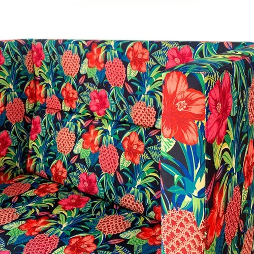 Close-up of a colorful floral patterned fabric with red hibiscus flowers, pink pineapples, green leaves, and blue background on a sofa.