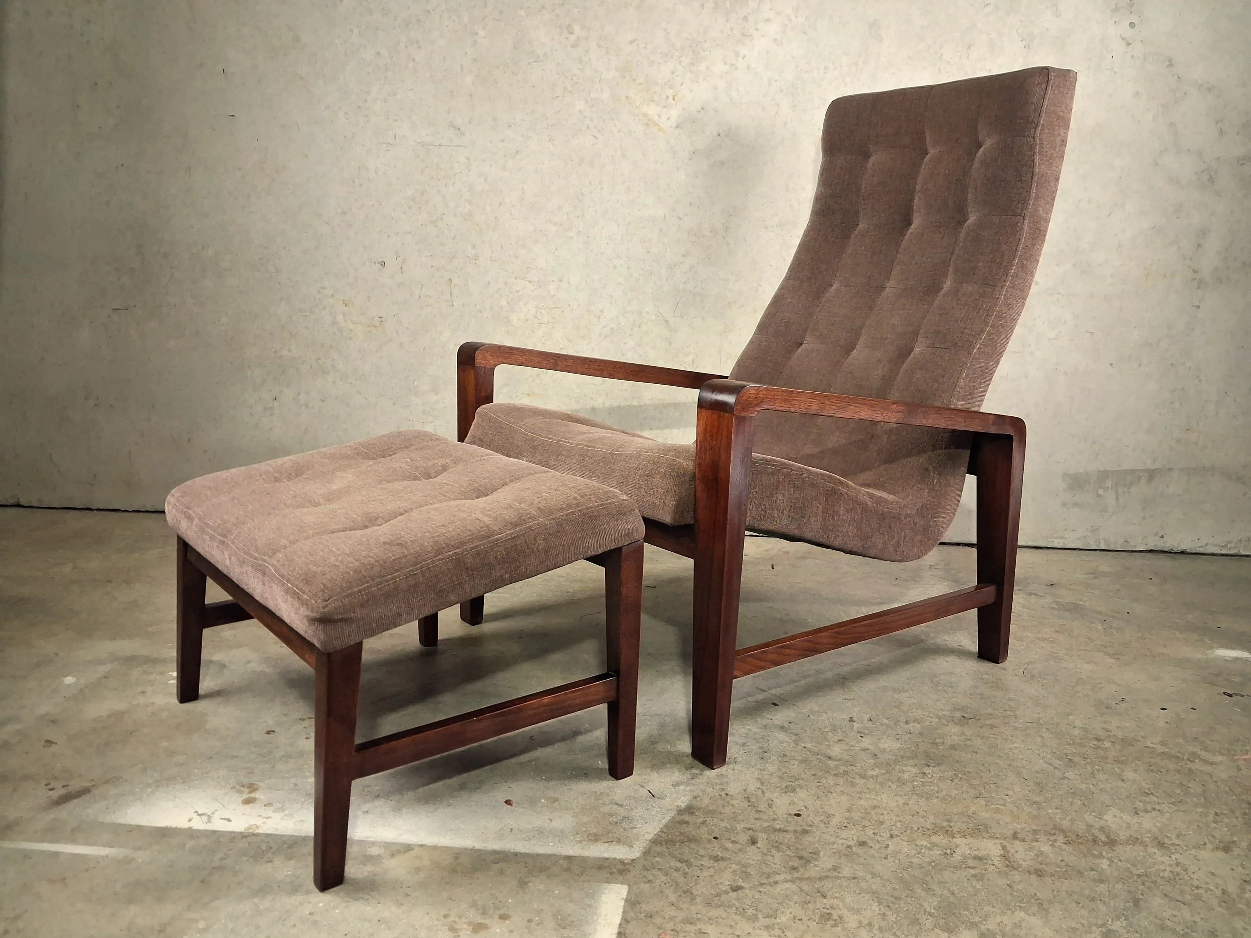 A mid-century modern armchair with a matching ottoman, both featuring wooden frames and beige upholstered cushions, placed on a concrete floor against a plain wall.