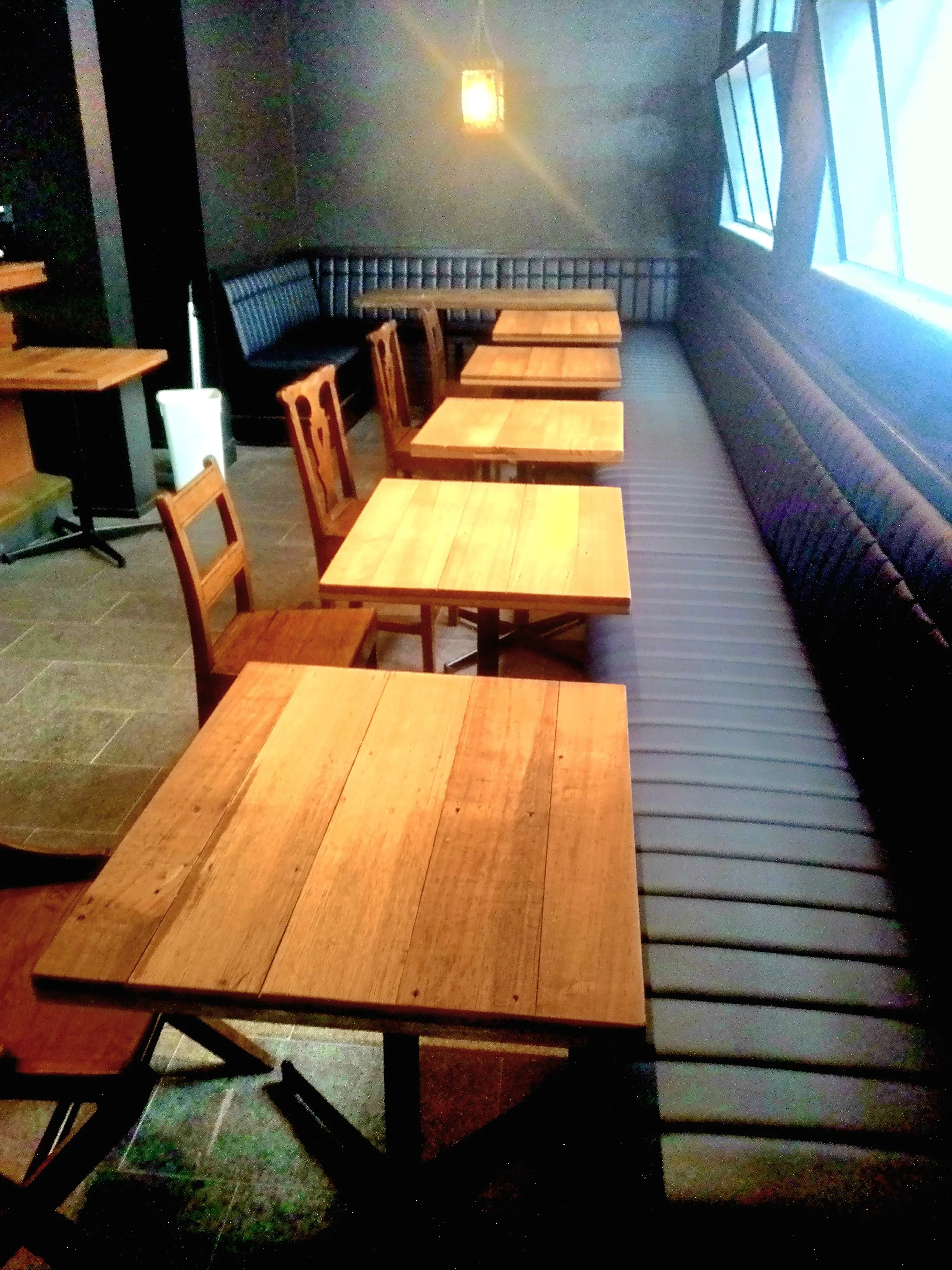 Empty dining area with wooden tables and chairs, black cushioned banquette seating along the wall, and a window with natural light.